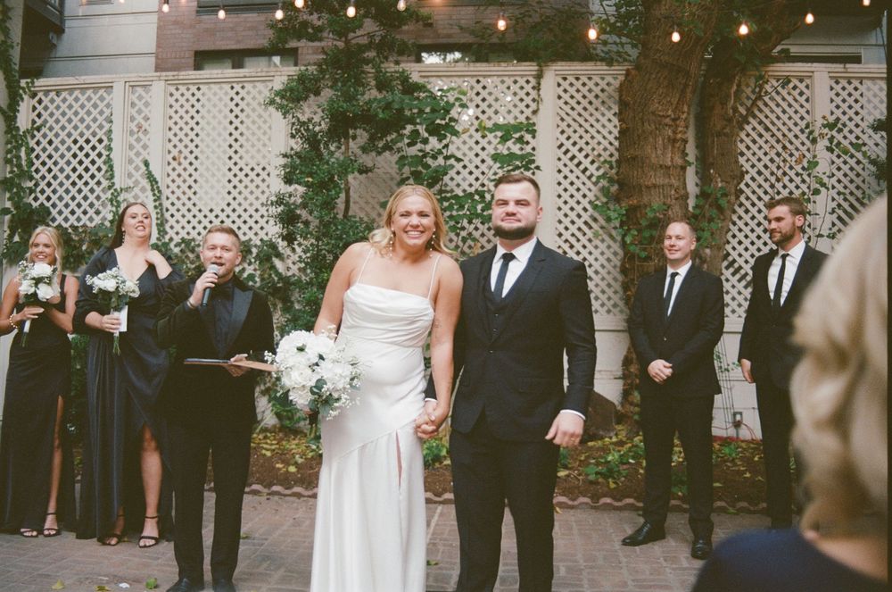 Wedding party poses together outdoors in front of white lattice backdrop during formal ceremony.