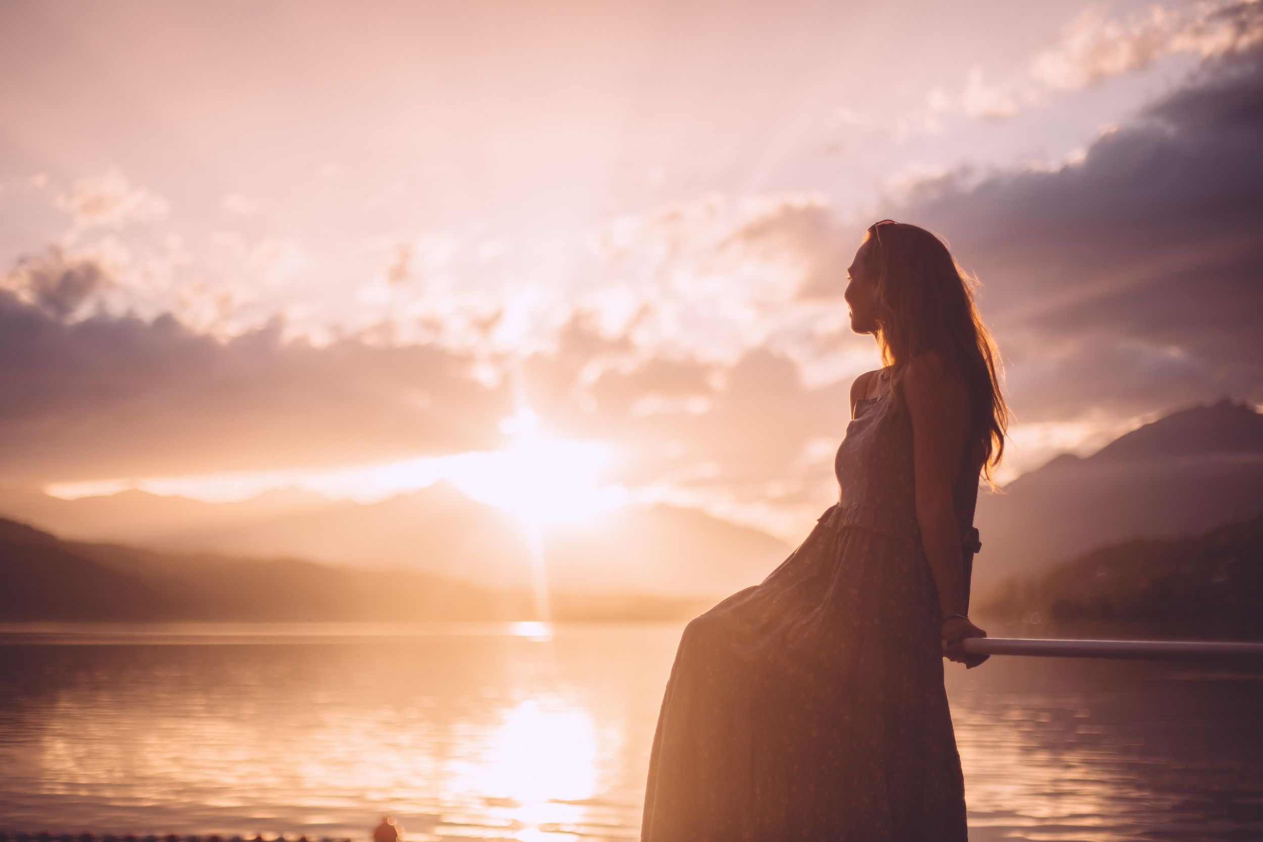 Silhouetted figure stands on beach at sunset with dramatic orange sky and mountains in background.