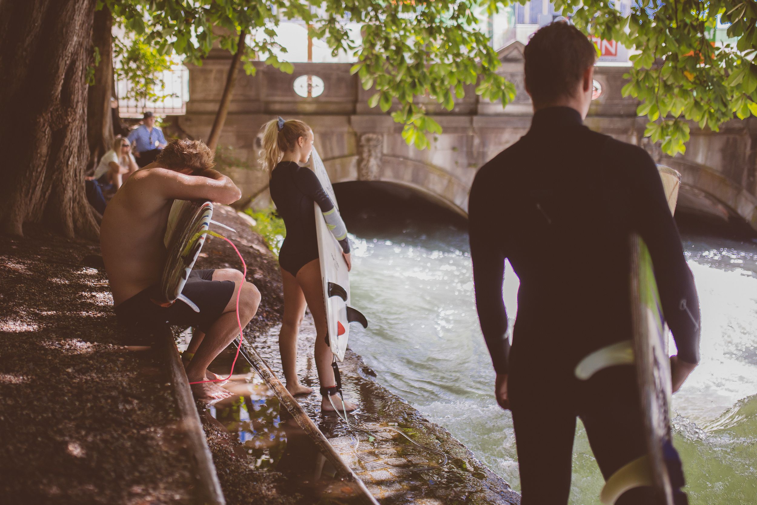 A group of people enjoy swimming and playing in a natural swimming hole surrounded by trees and rocks on a sunny day.