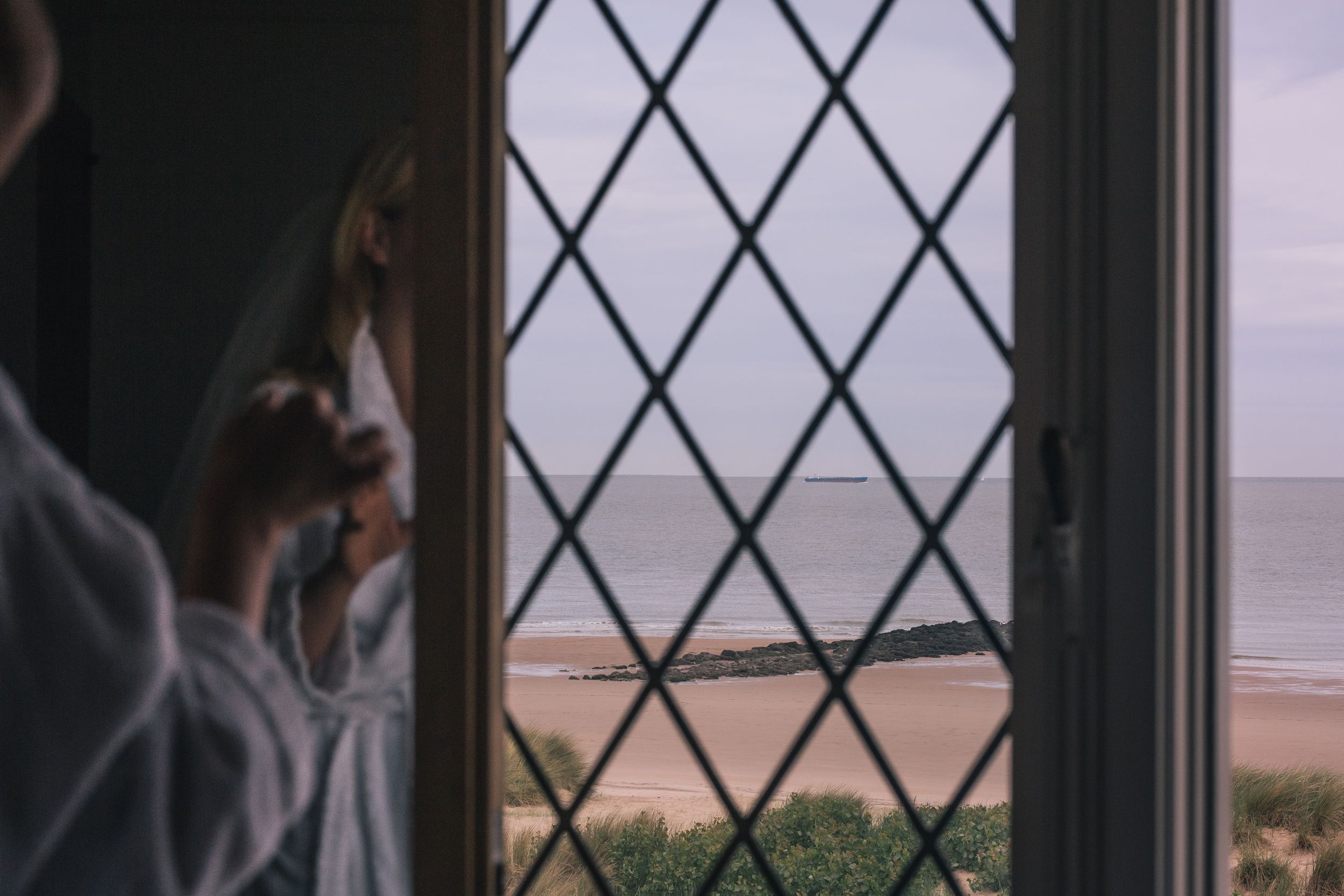 Person sitting by a window with diamond-patterned glass panes overlooking a coastal landscape at sunset.