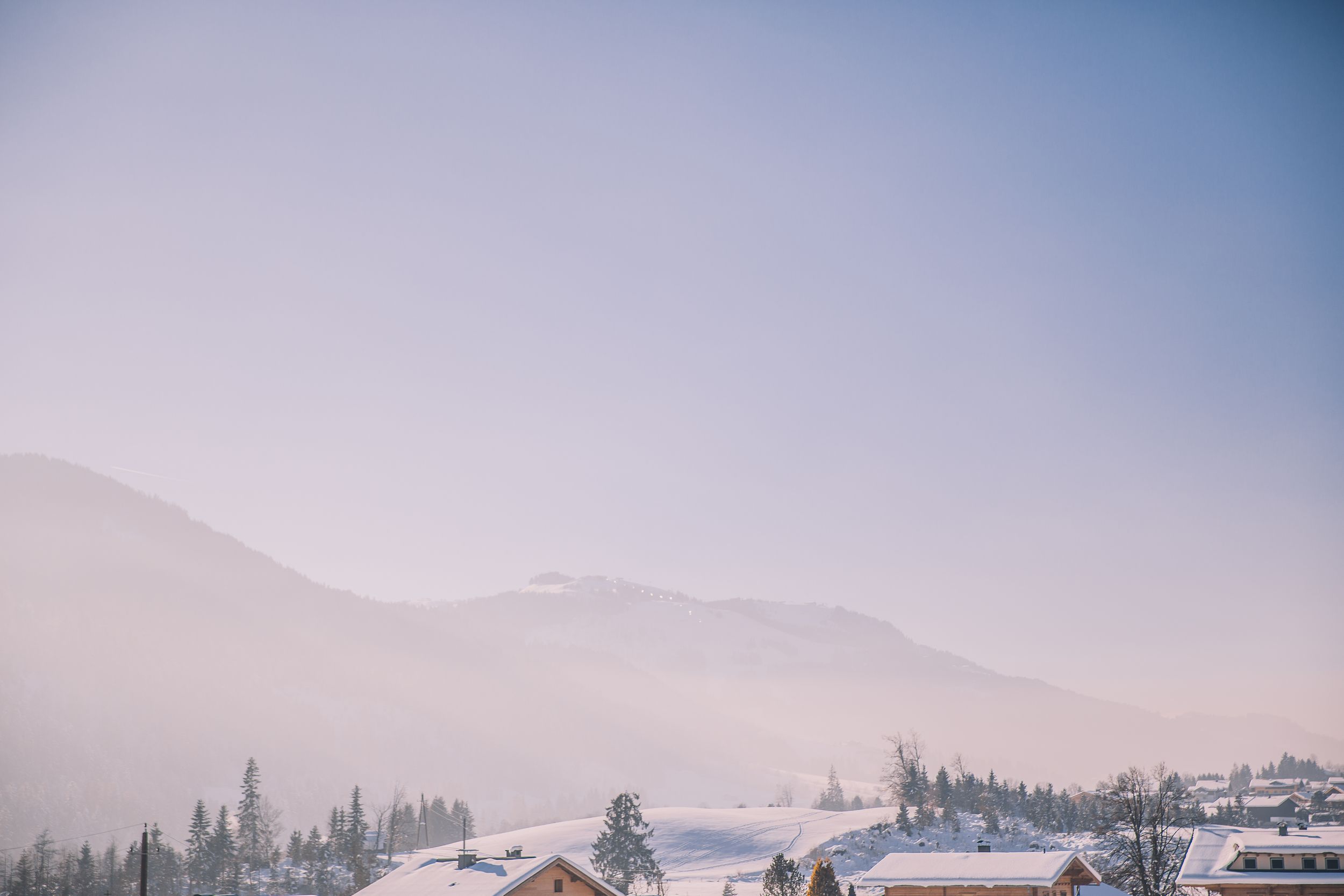 Scenic mountain landscape with evergreen trees and snow-capped peaks against a misty blue sky at sunset.