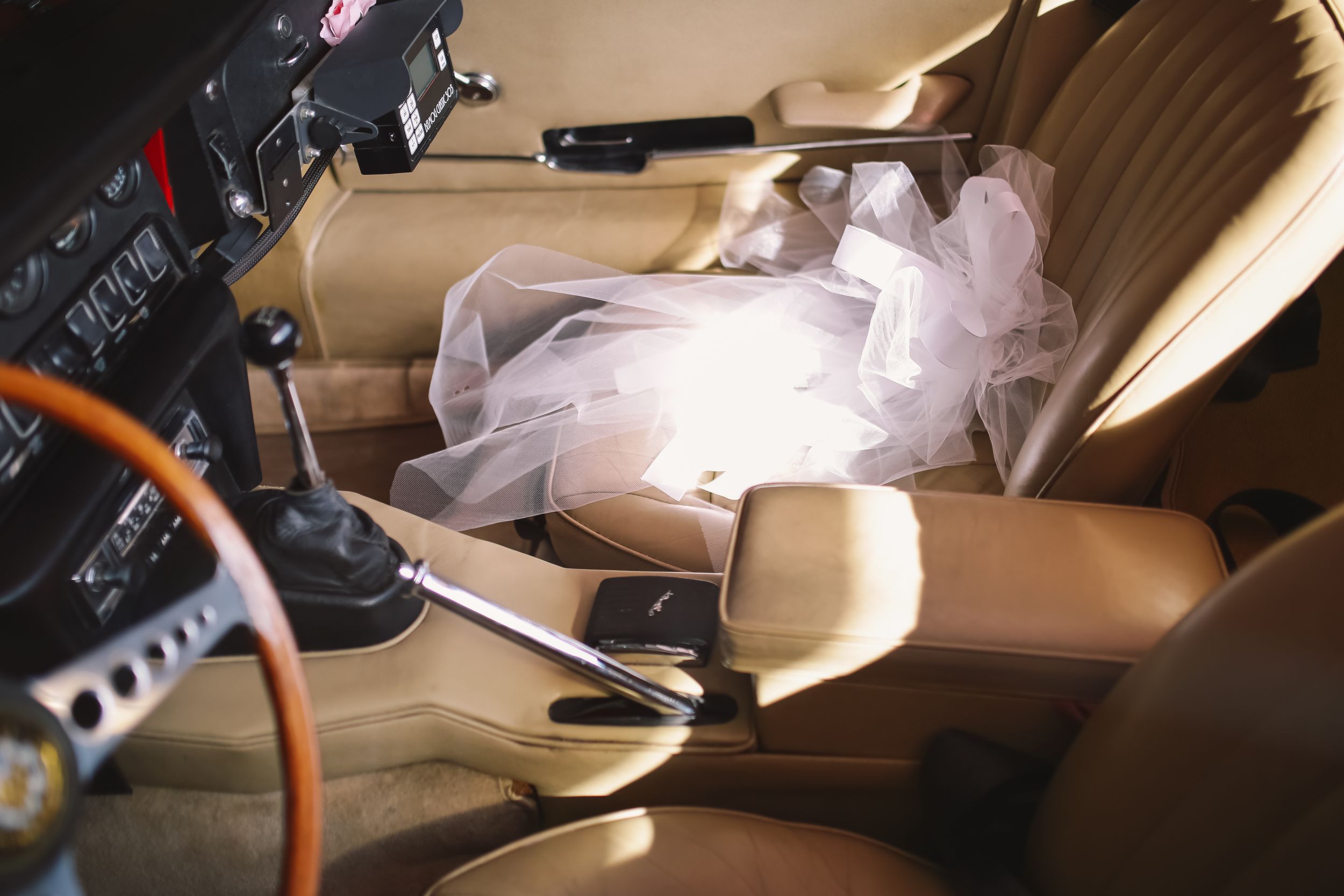 A wedding dress lays across tan leather car seats next to a wooden steering wheel.