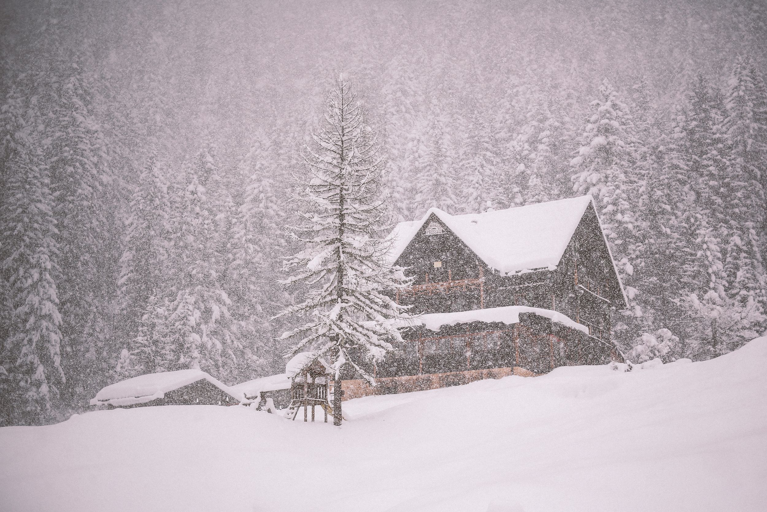 Snow falls heavily on rustic wooden cabins and evergreen trees in a serene winter mountain landscape.