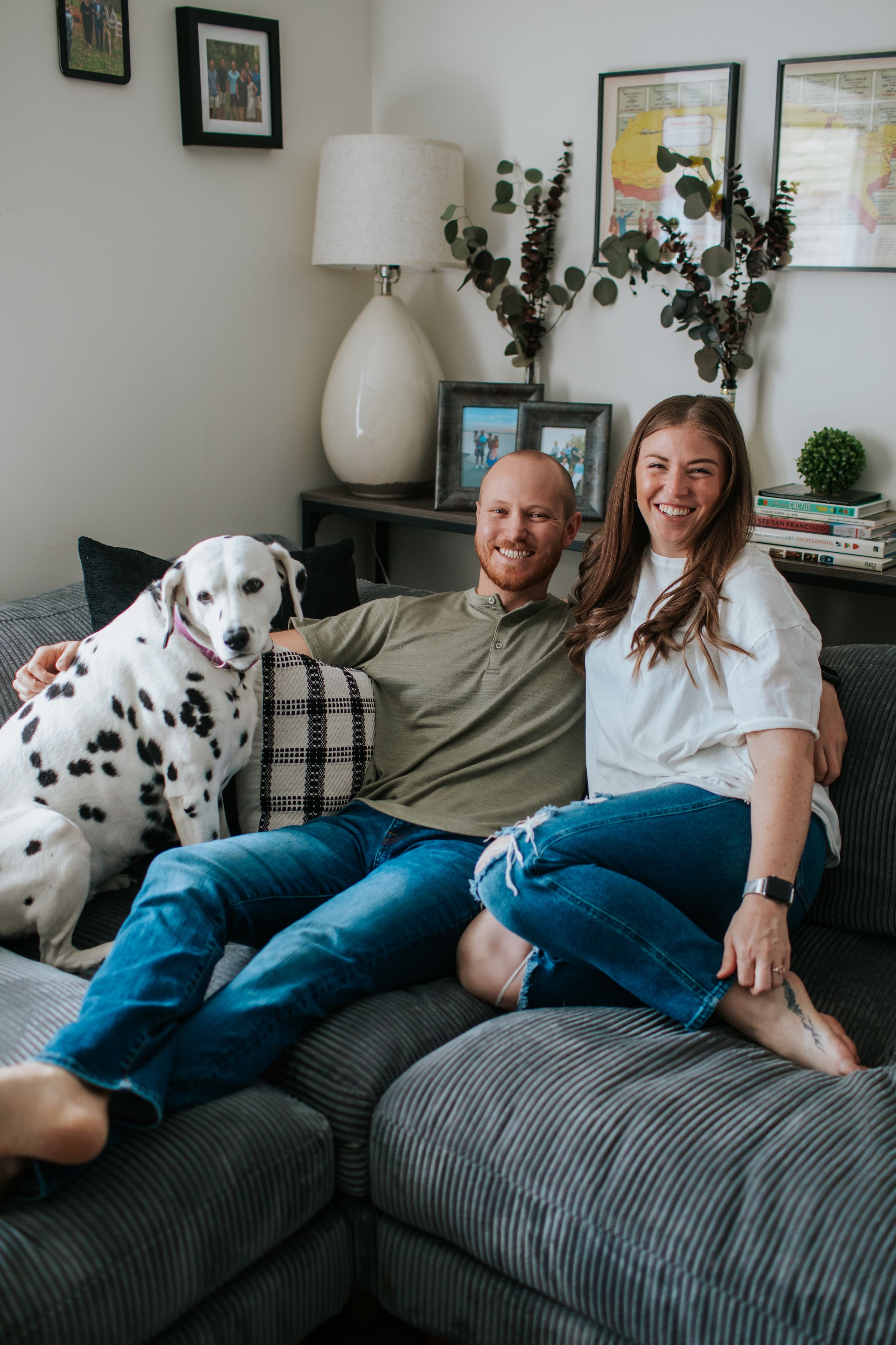 A couple relaxes on a gray couch with their spotted Dalmatian dog in a cozy living room with lamp and wall art.