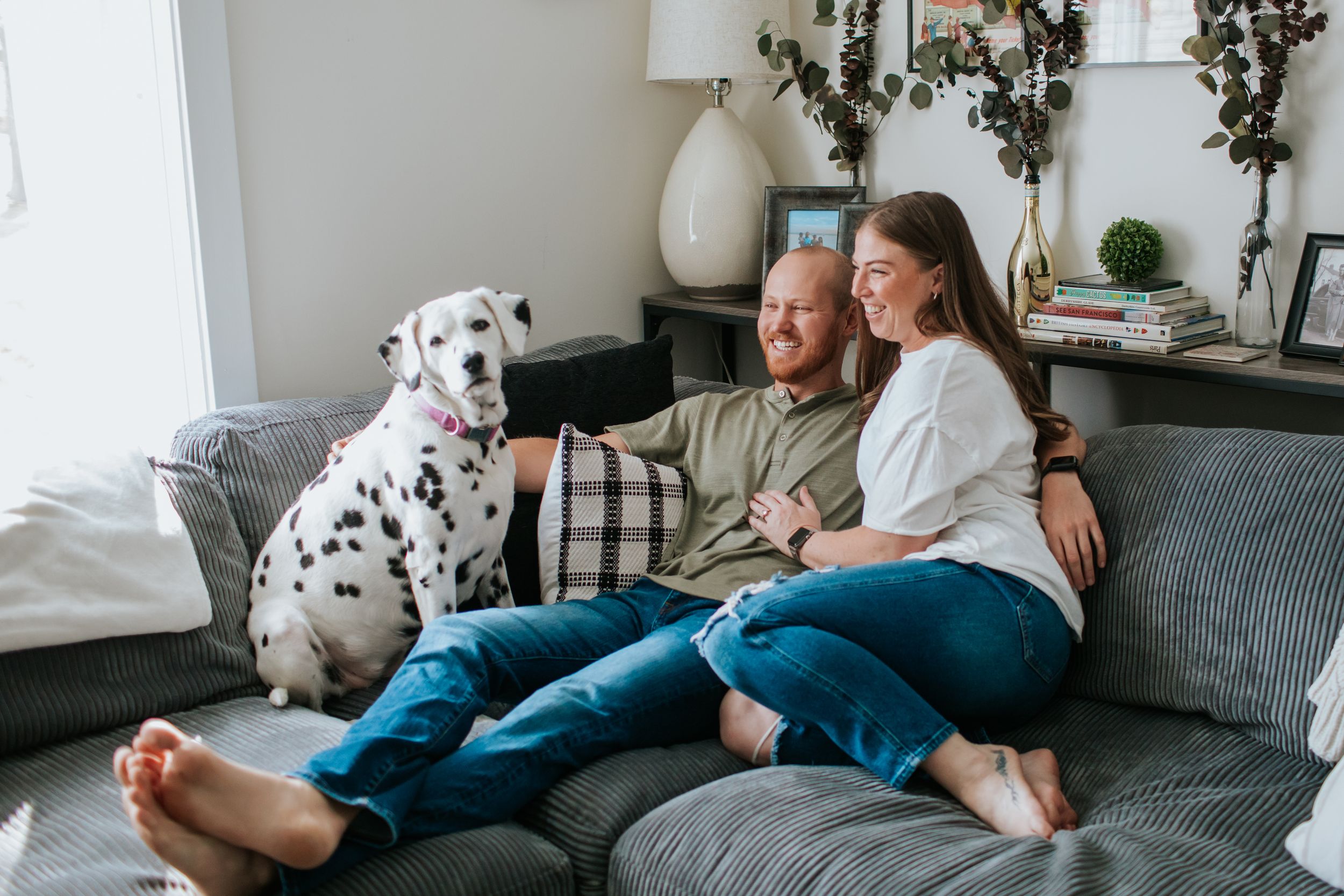 A person and dog relax together on a grey couch in a bright living room with natural light streaming in.