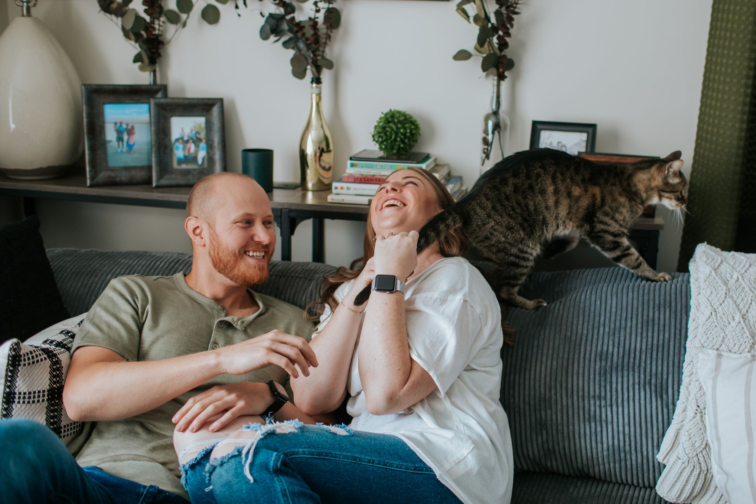 A couple relaxes together on a gray couch with greenery hanging on the wall behind them.