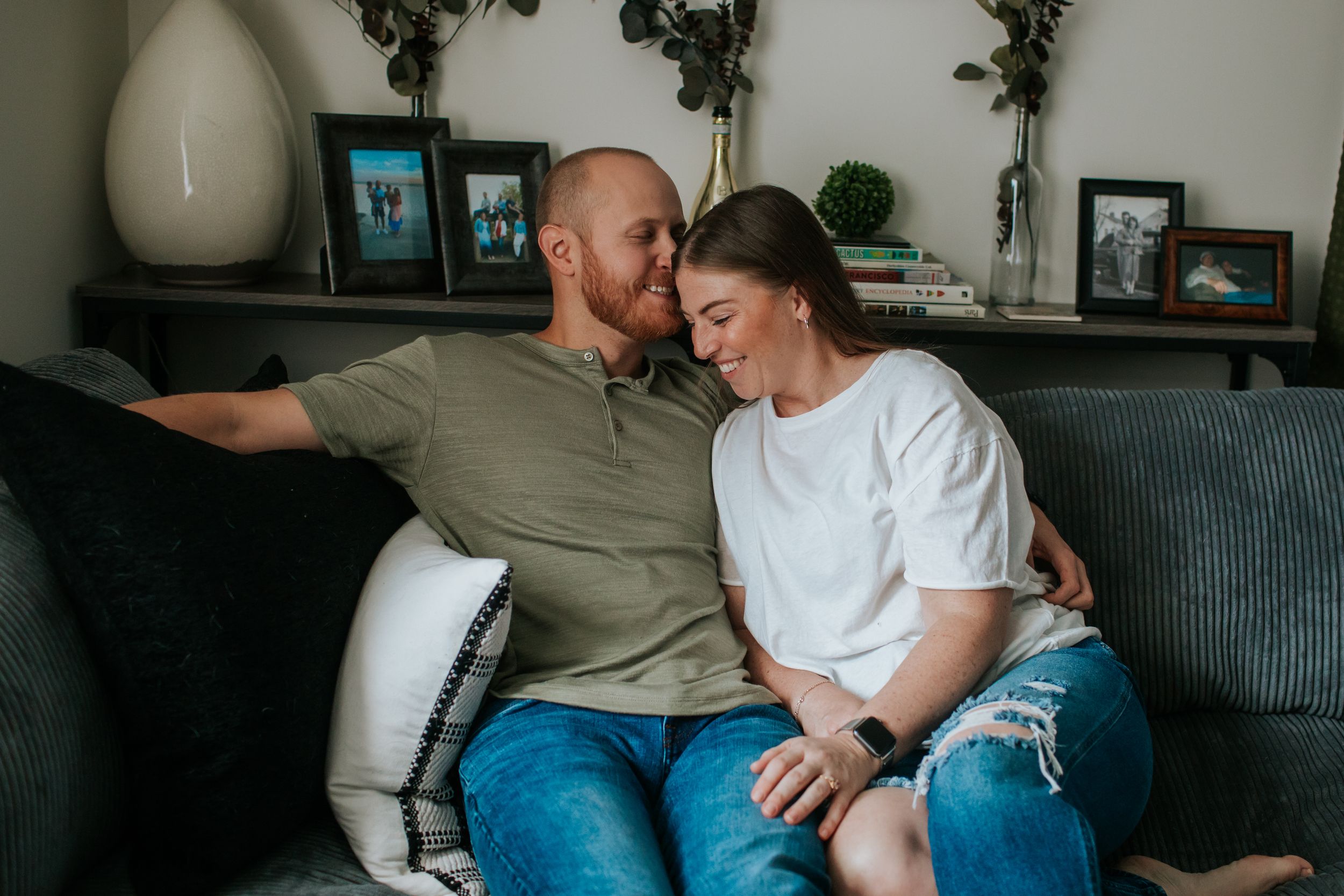A couple sits together on a grey couch in a cozy living room setting, sharing an intimate moment.