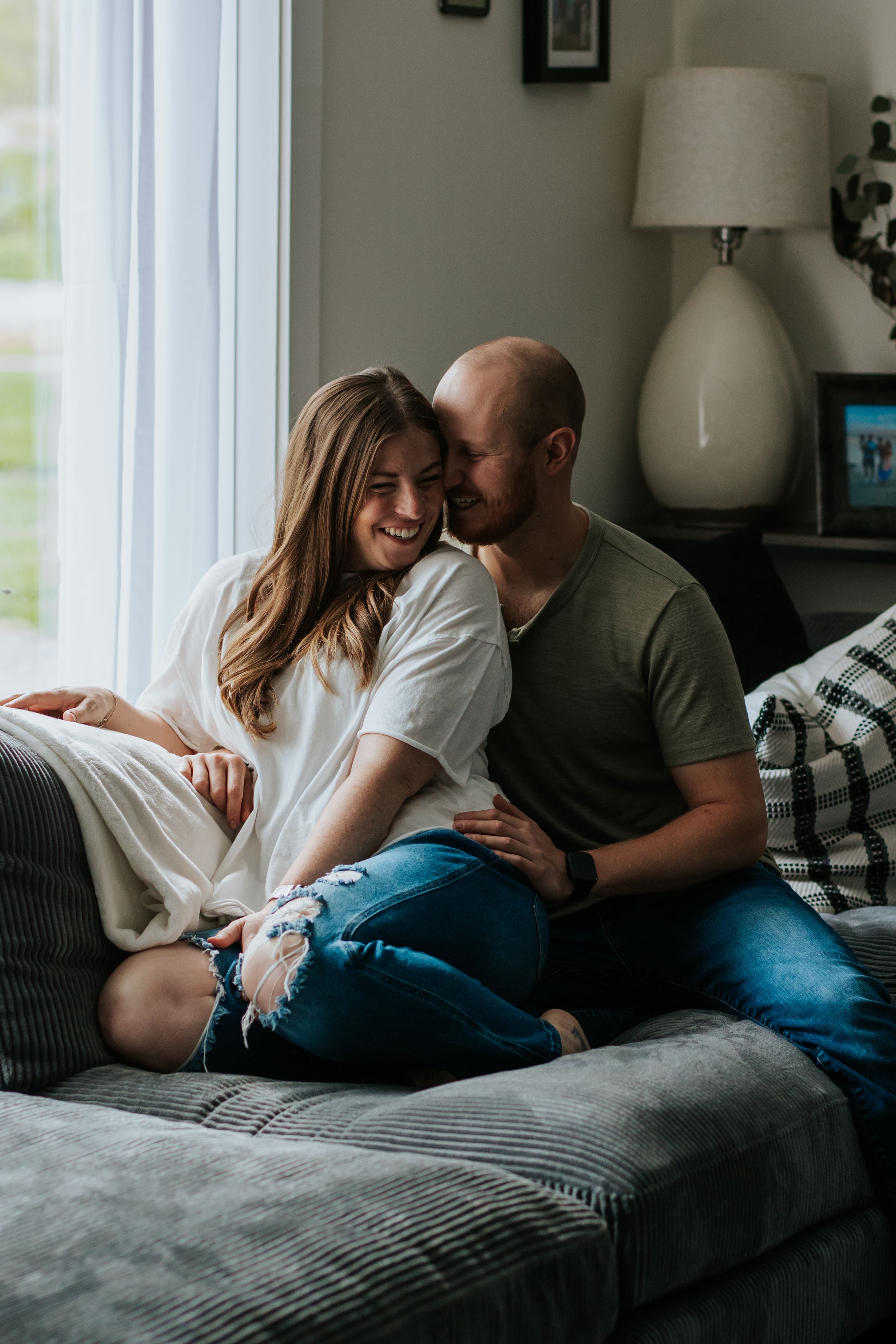 A couple shares an intimate moment while relaxing together on a gray couch in a naturally lit room.
