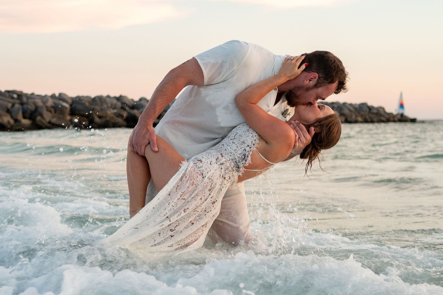Romantic couple in white outfits embracing and dancing on a sandy beach at sunset by the ocean waves.