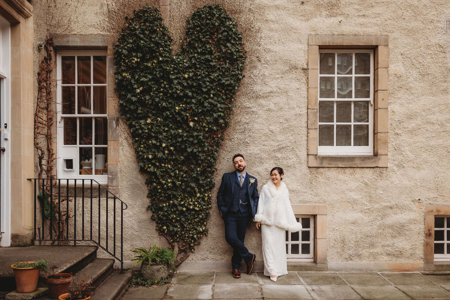 A couple stands in front of a historic stone building with ivy-covered walls and windows.