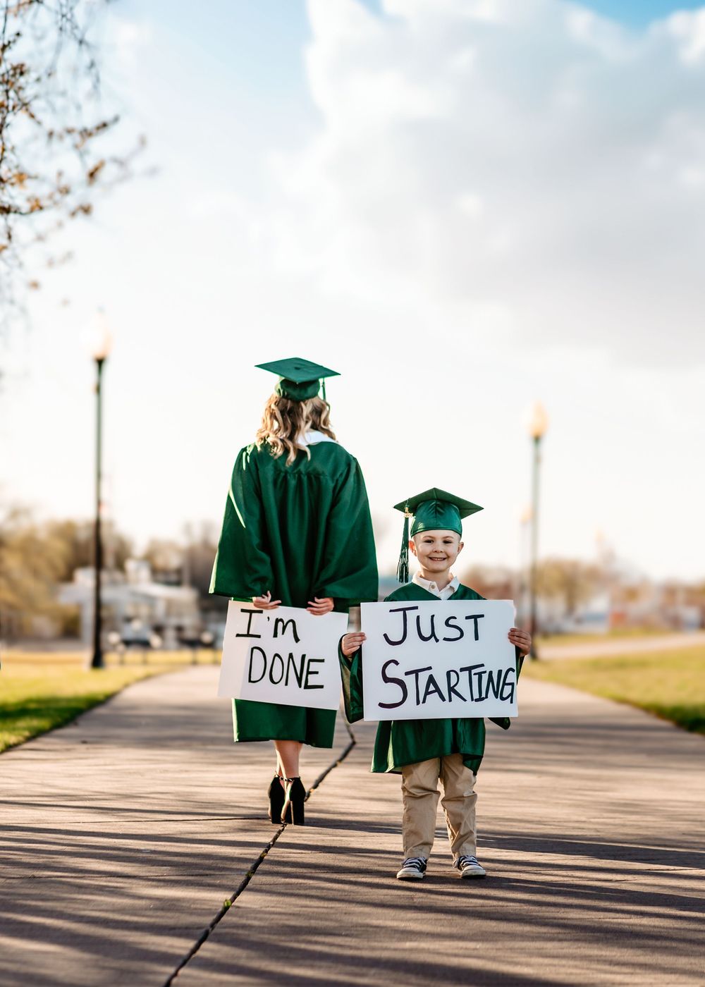 Two graduates in green caps and gowns hold signs reading 'I'm Done' and 'Just Starting' while walking down a path.