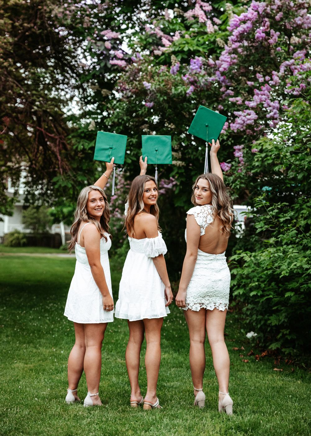 Three women in white dresses hold up green signs while standing on grass near blooming lilac bushes in a garden setting.