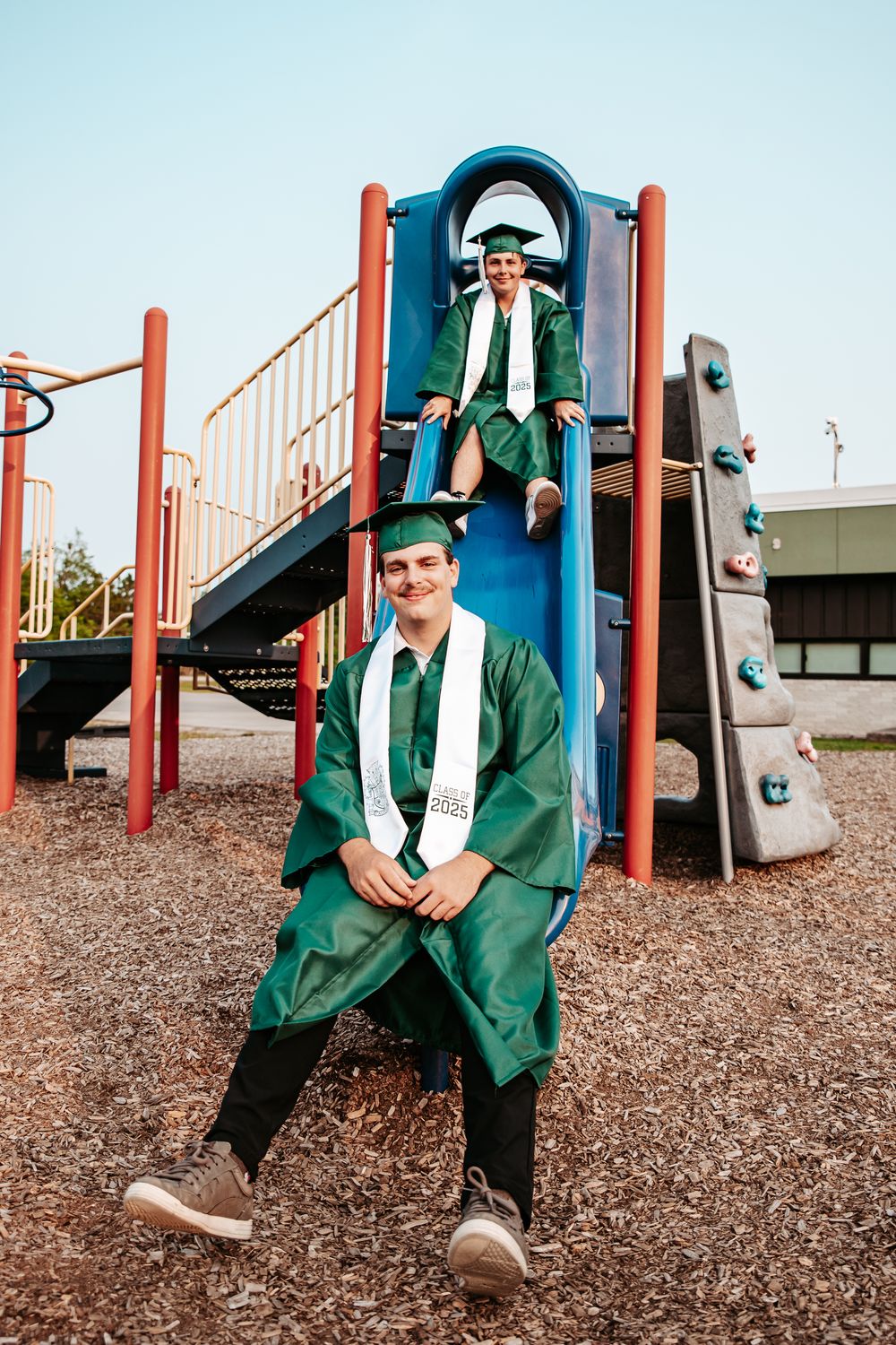 Graduates in green caps and gowns pose playfully on a playground slide during a celebratory photoshoot.