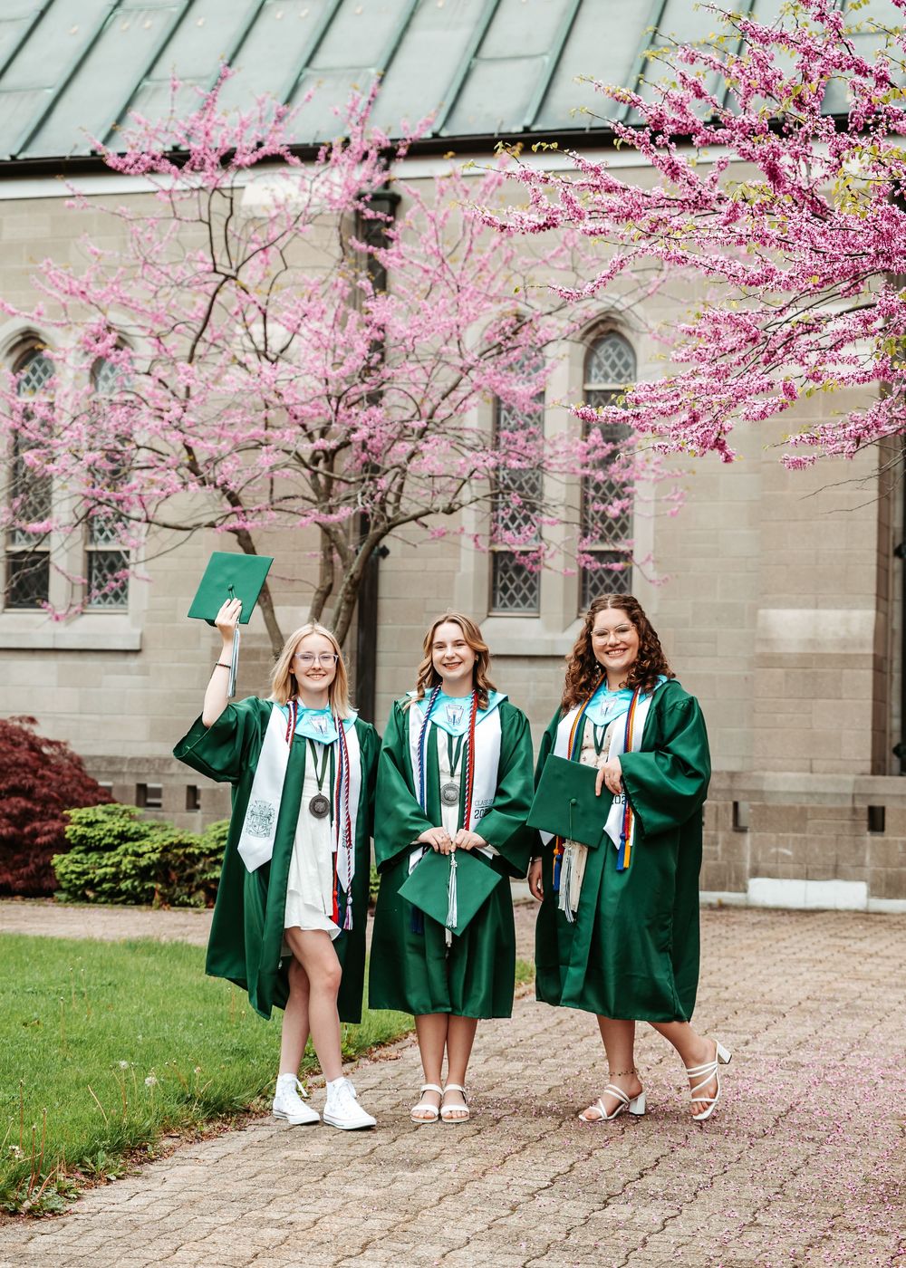 Three graduates in emerald green robes celebrate under blooming cherry blossoms by a stone church with stained glass windows.