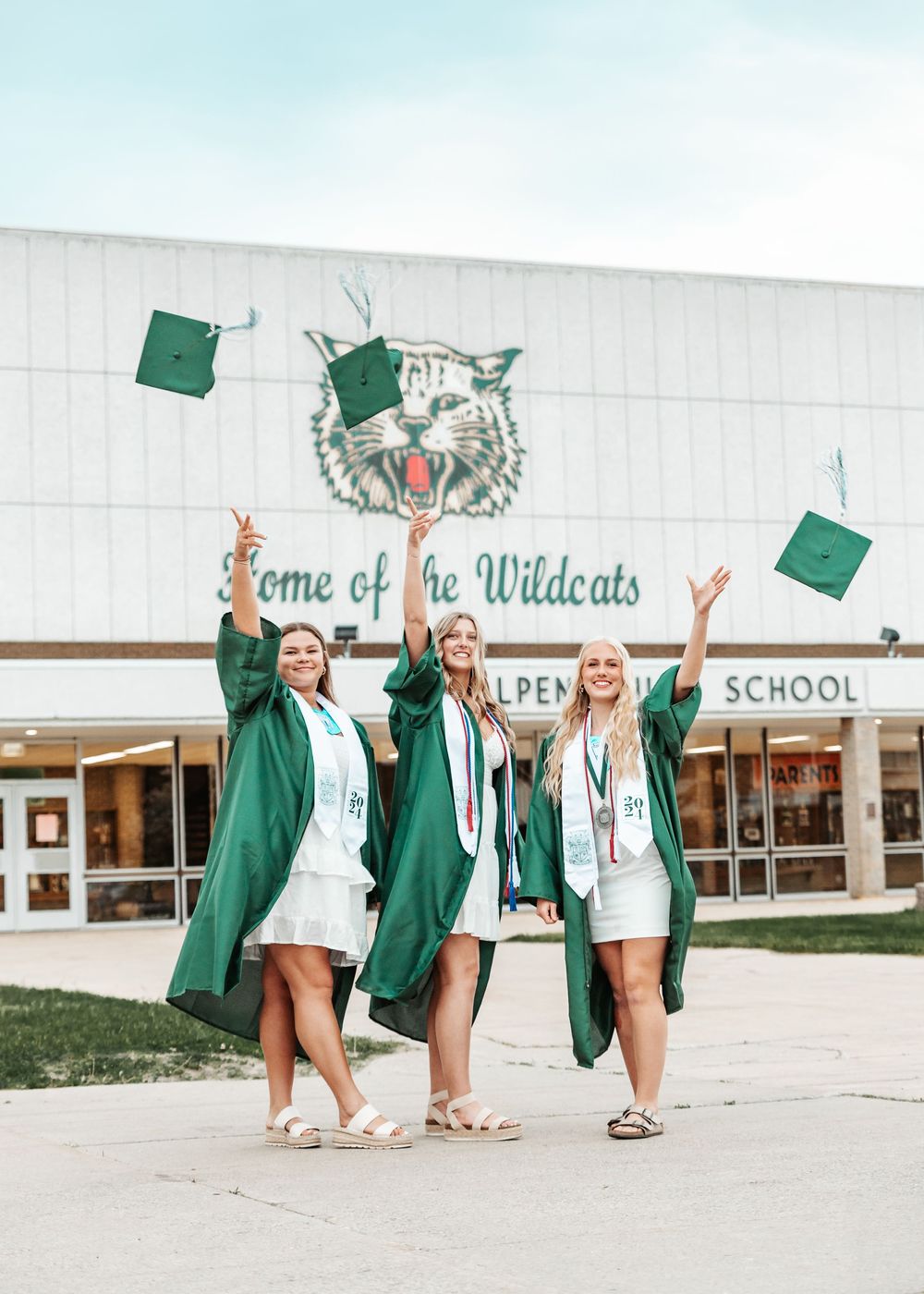 Graduates in green caps and gowns toss their mortarboards in celebration outside their school building.