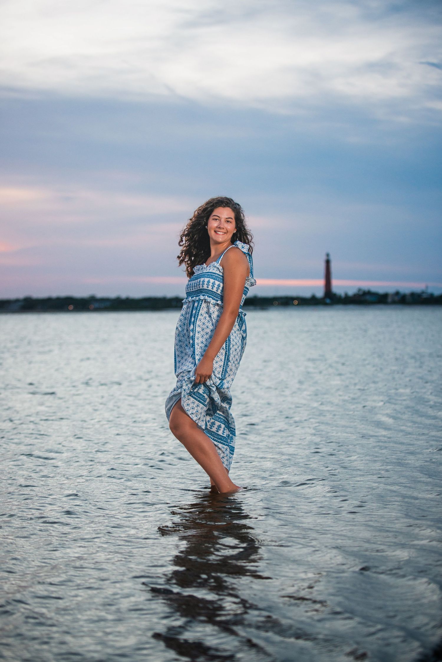 Woman in blue dress plays in water at sunset with lighthouse in background.