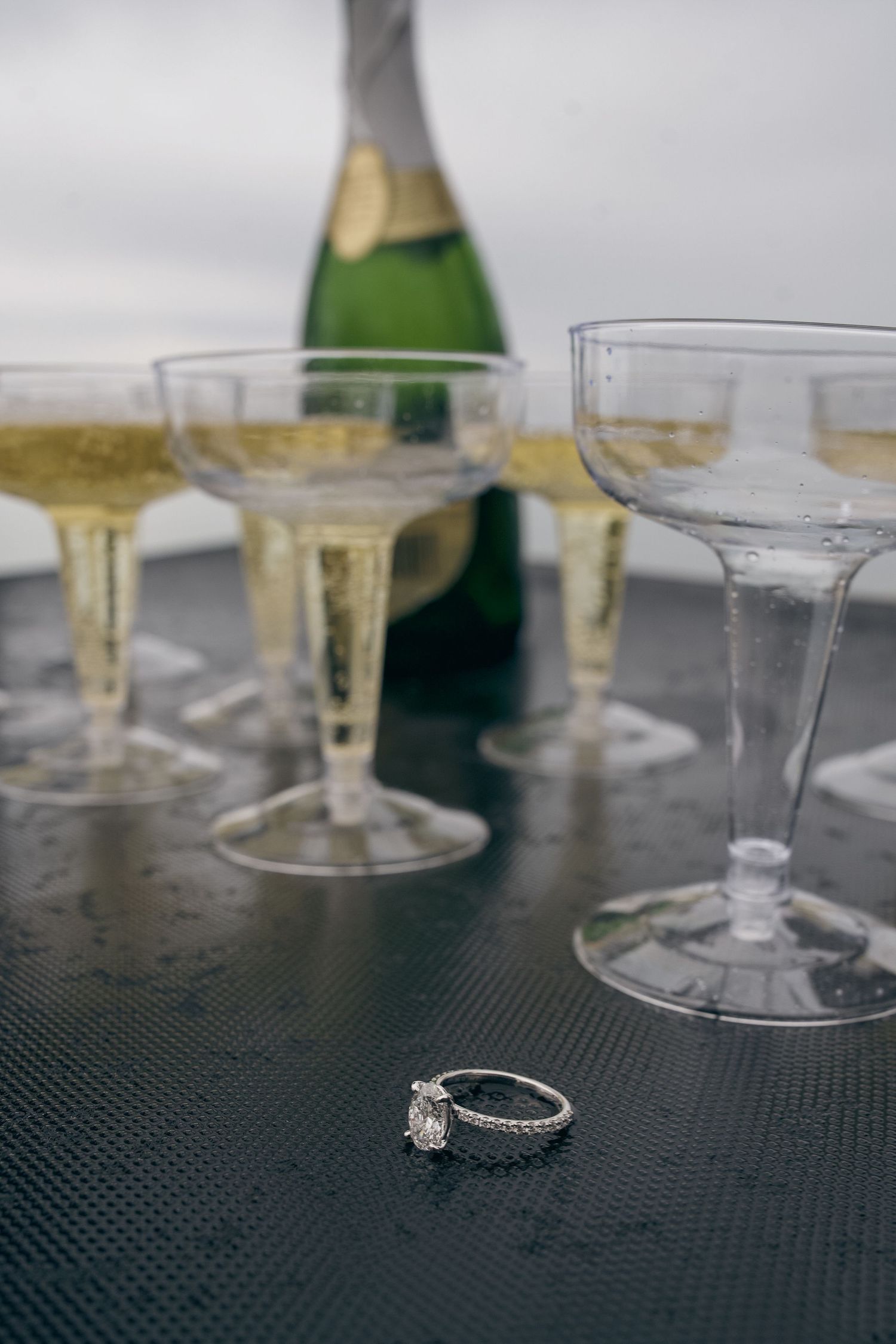 Elegant diamond engagement ring sits on black textured surface with champagne glasses blurred in background.