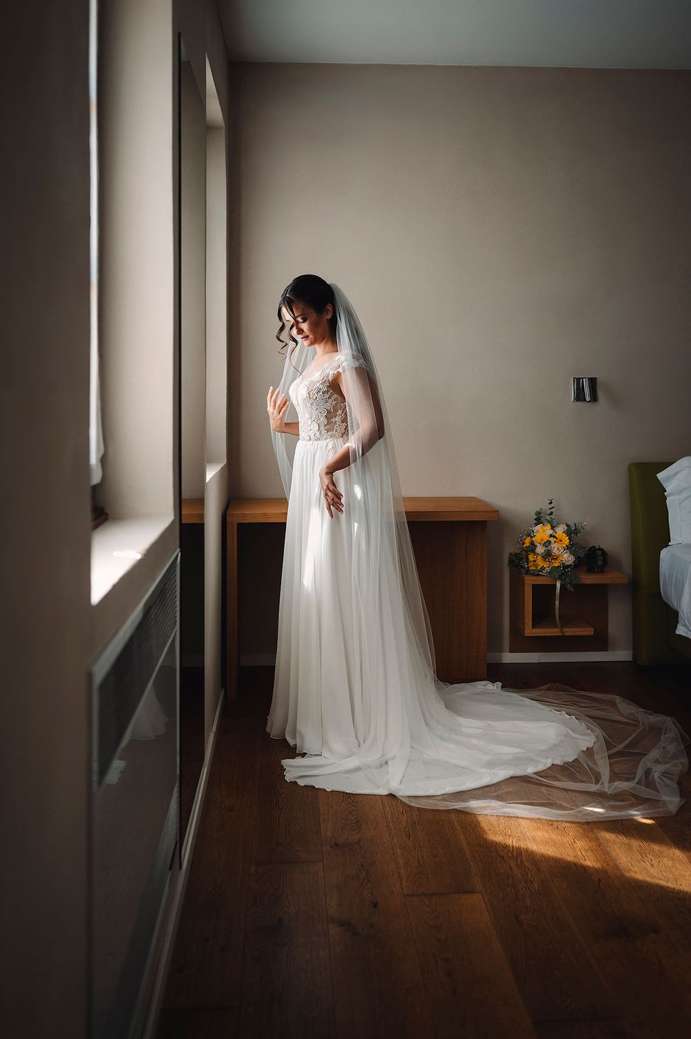 Una sposa con un abito bianco e un velo è in piedi vicino alle finestre illuminate dal sole in un ambiente interno sereno.