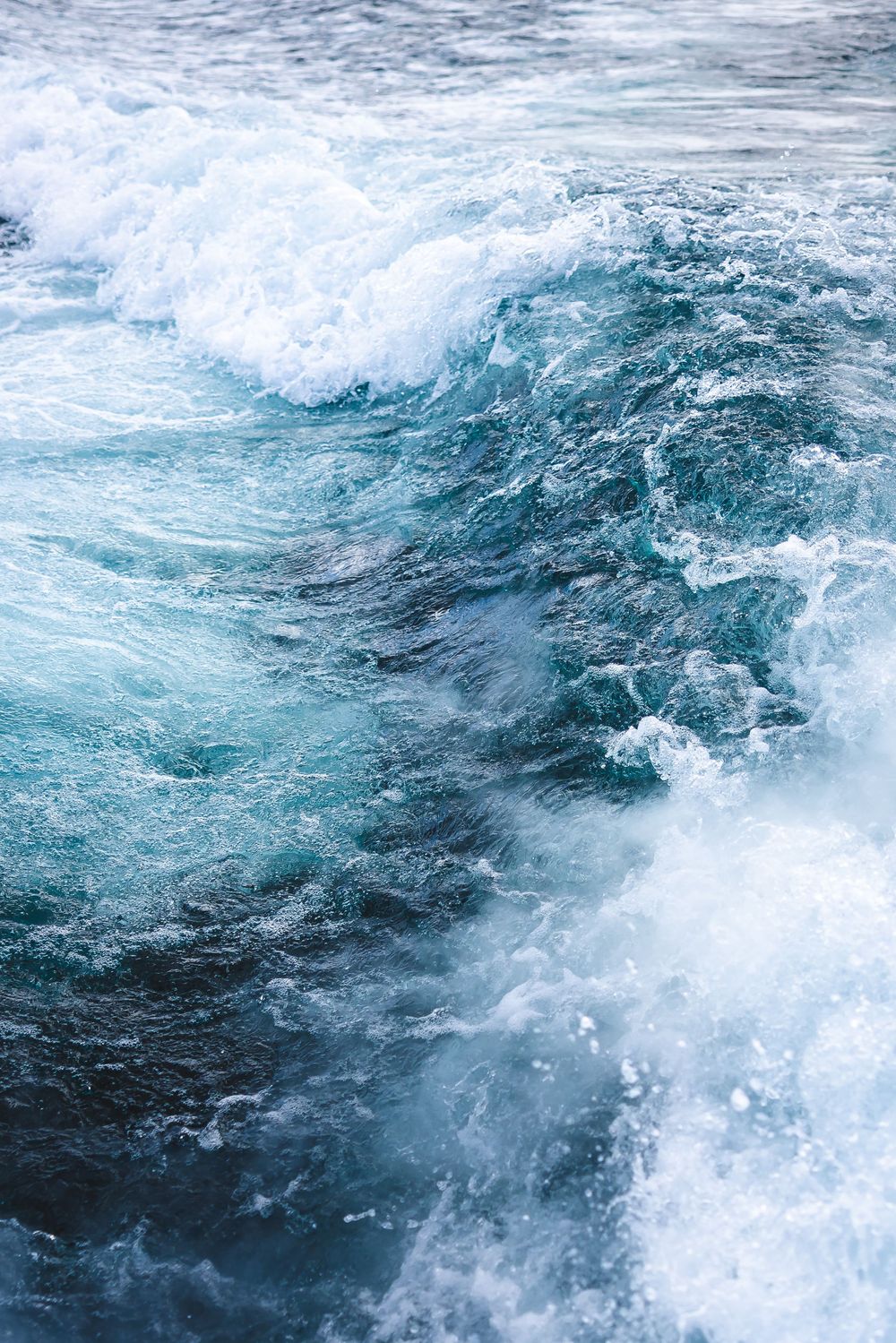 Turbulent ocean waves create a dramatic swirl of blue-green water and white foam against a dark surface.