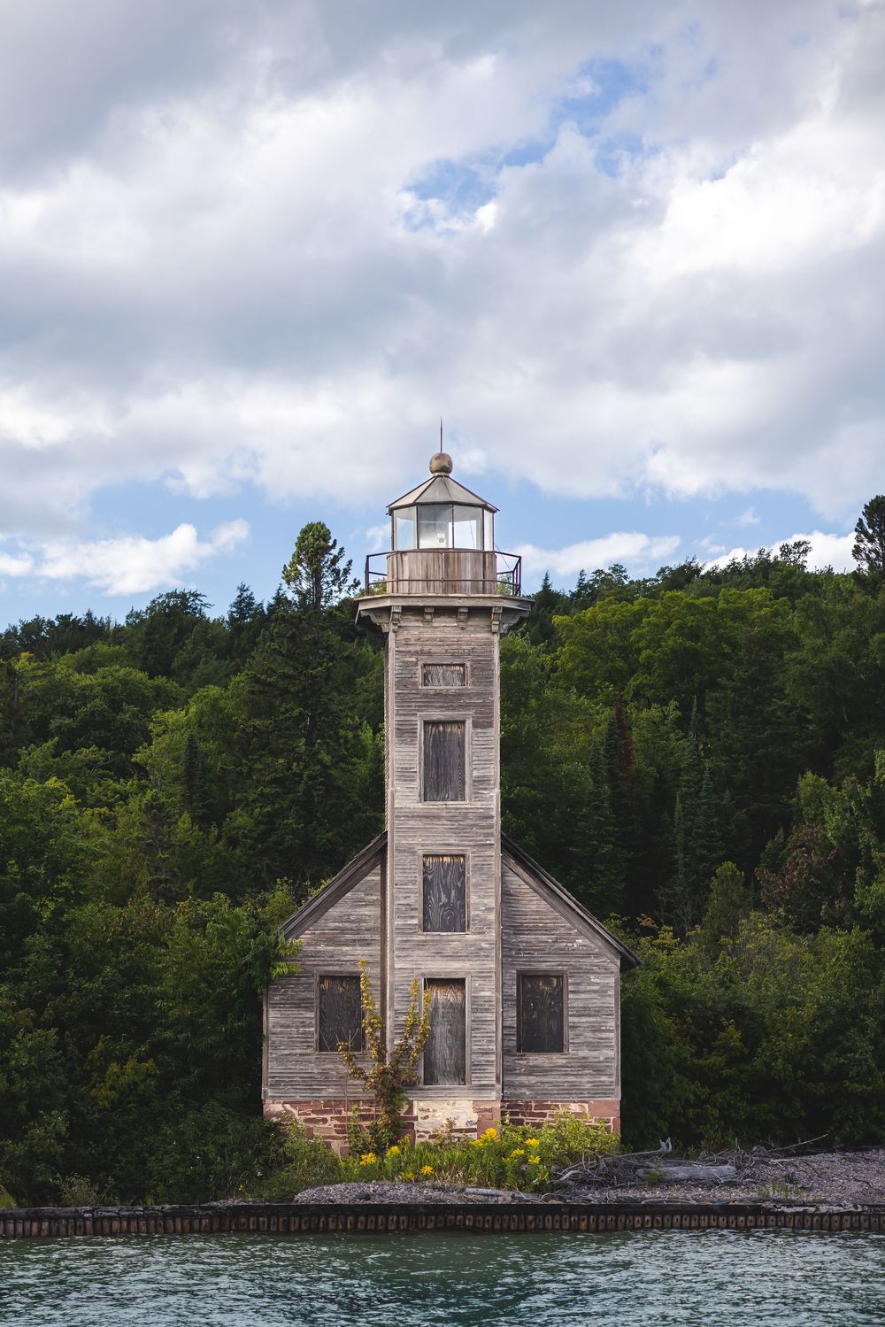 Historic wooden lighthouse with weathered siding stands on rocky shoreline against forested backdrop and cloudy sky.