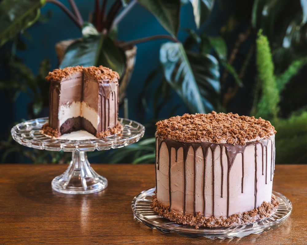 A chocolate layer cake with dripping ganache sits on a glass cake stand against a dark background.