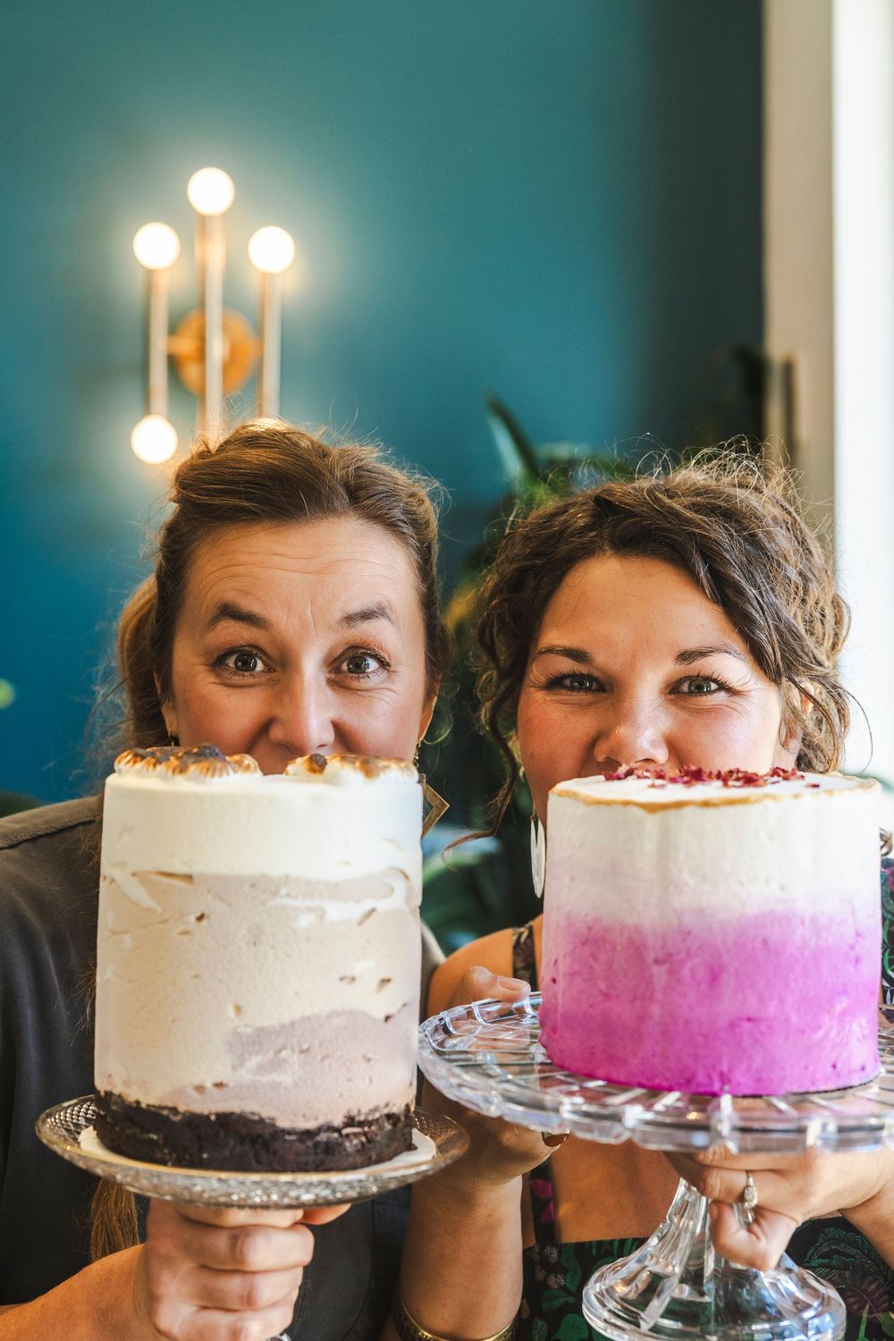 Two cakes with white frosting and pink ombre decoration are displayed on cake stands against a teal blue wall.