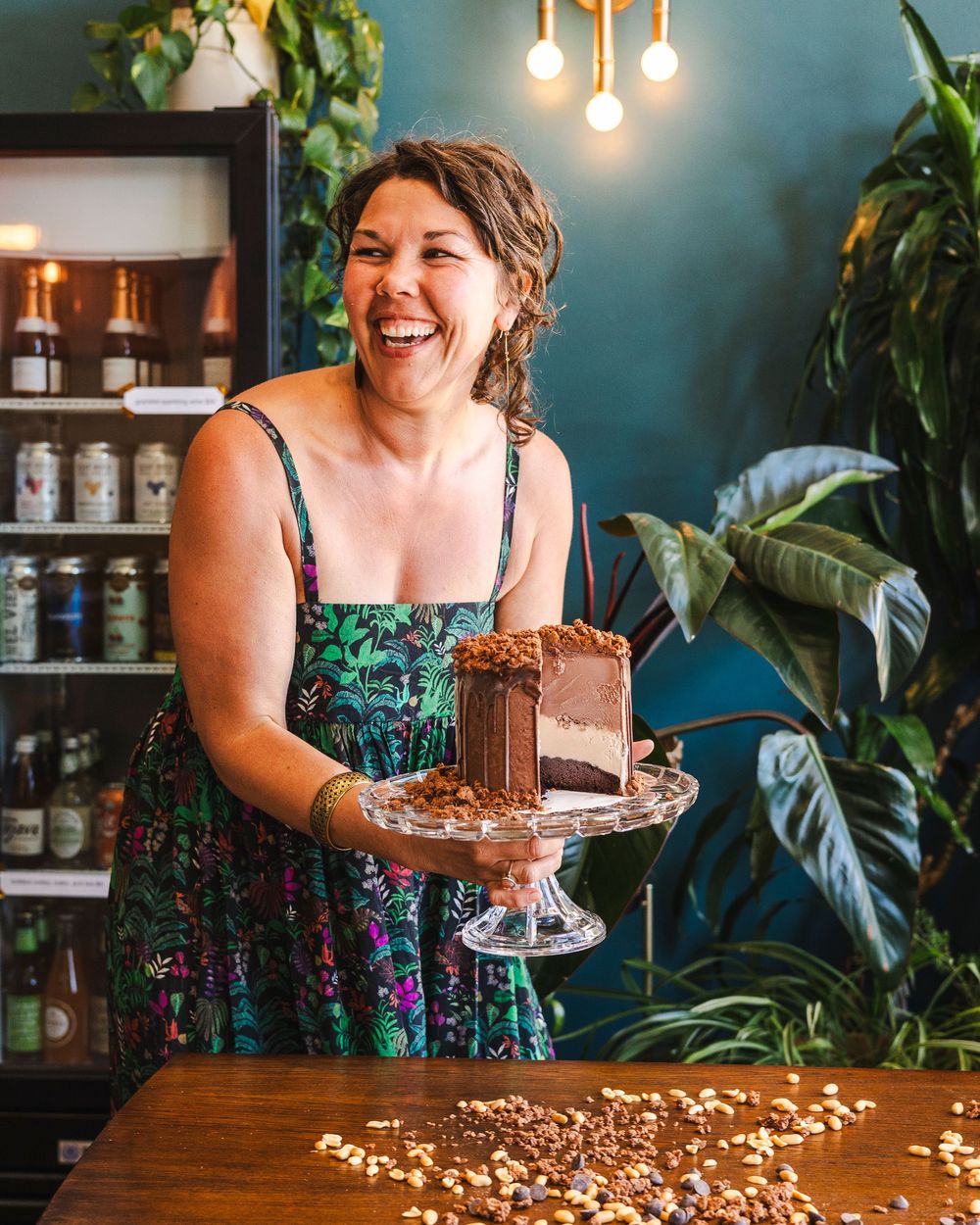 A person in a floral dress serves chocolate cake slices at a rustic wooden counter with hanging pendant lights and plants.