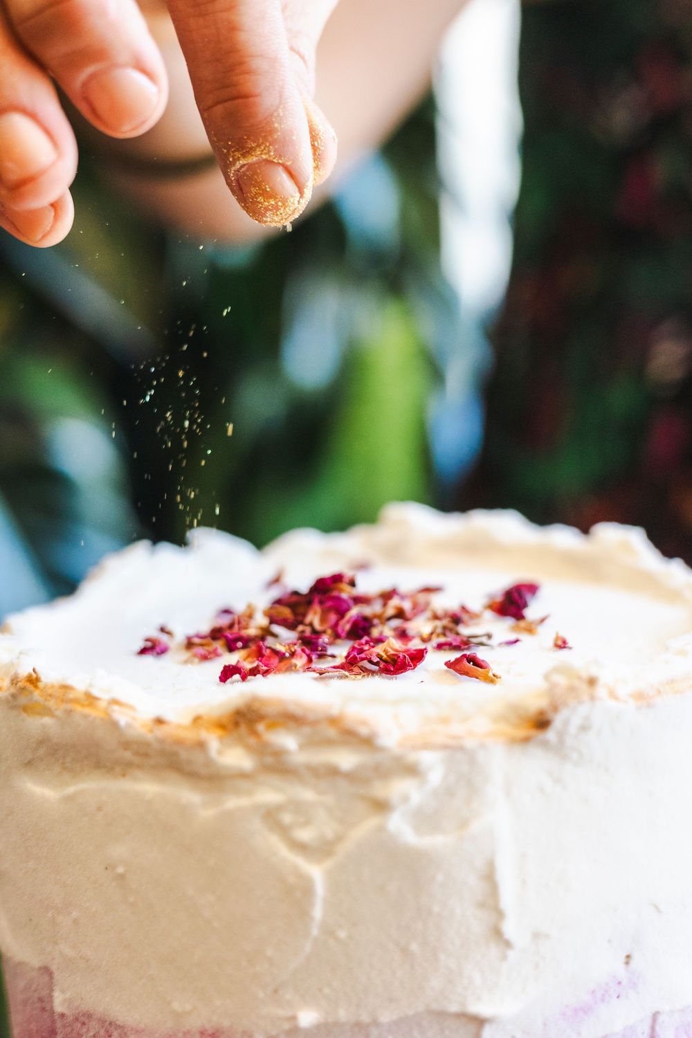 A hand sprinkles garnish over a white frosted dessert topped with red berries.
