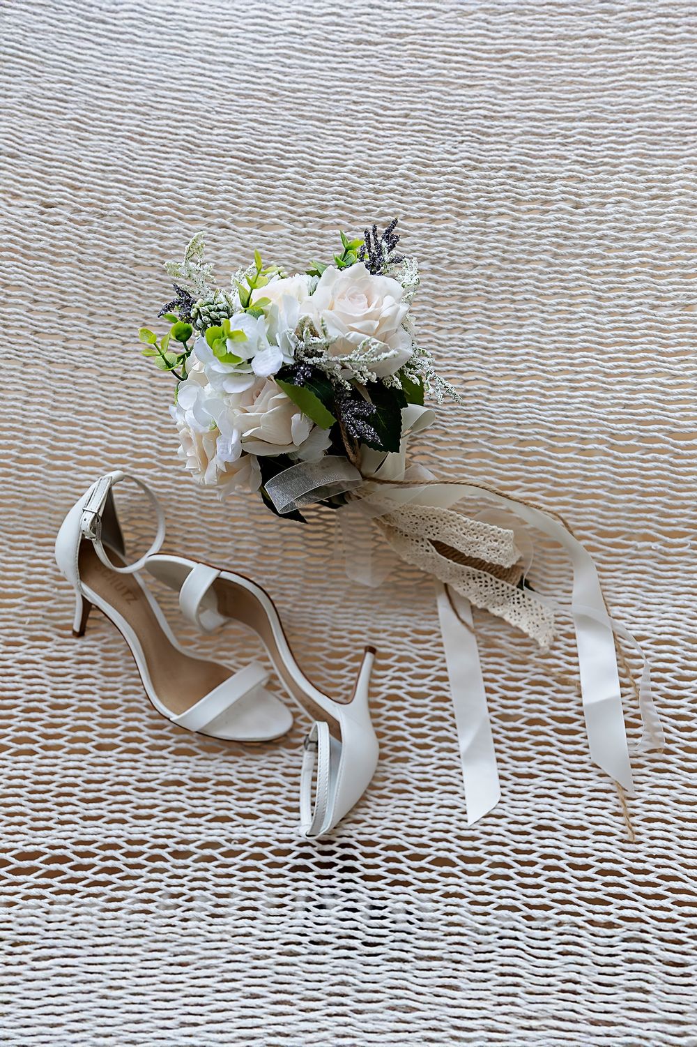 White strappy bridal sandals with floral decorations arranged on a textured beige surface.