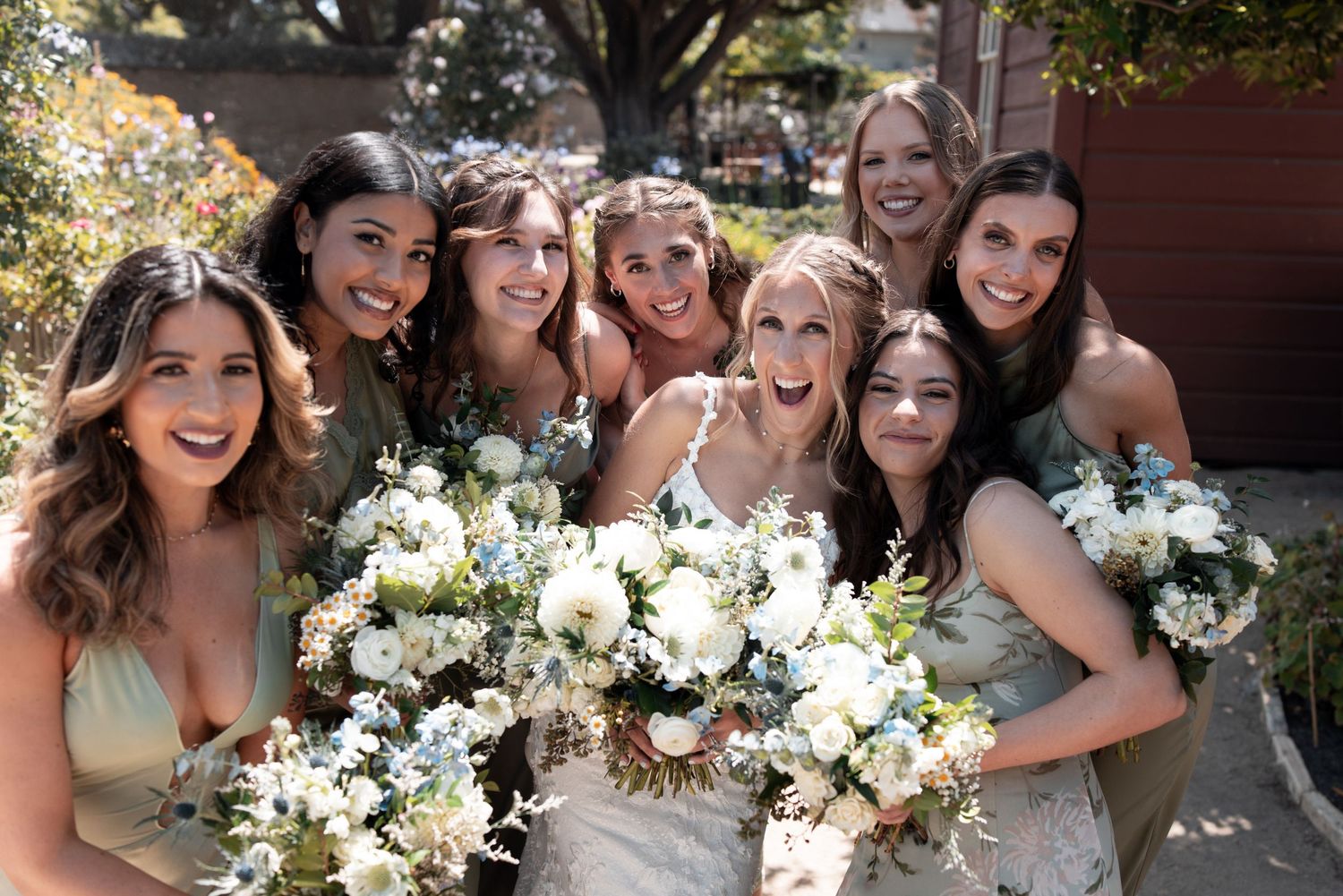 A group of bridesmaids in matching dresses hold white floral bouquets while posing together outdoors.