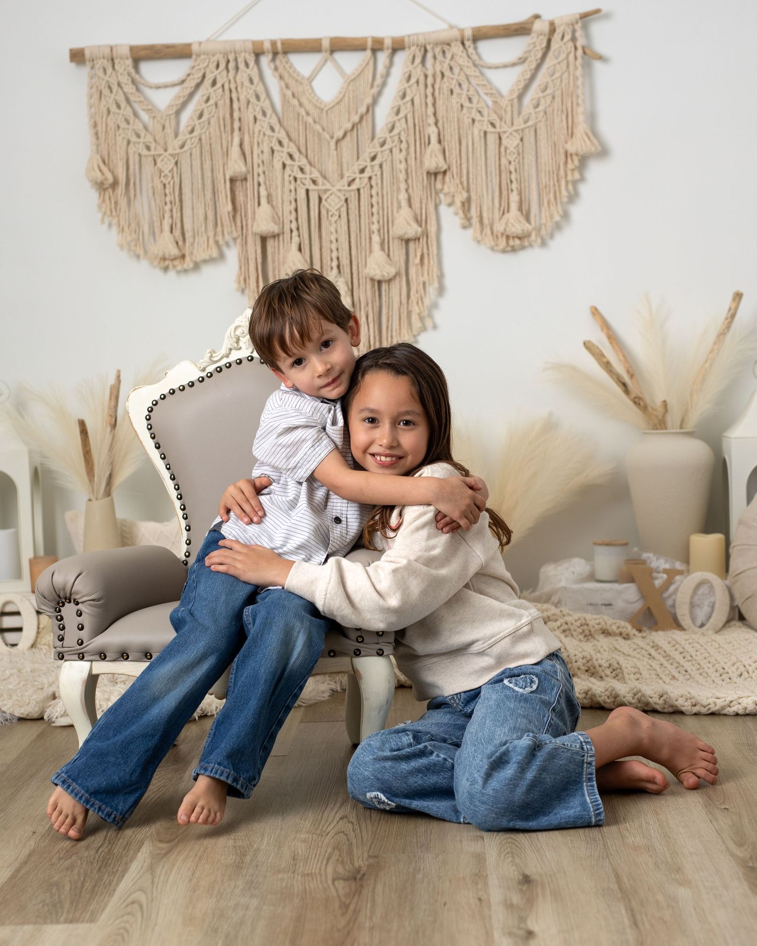 Two children in jeans and neutral sweaters play and laugh together on a floor with macrame wall hangings in the background.
