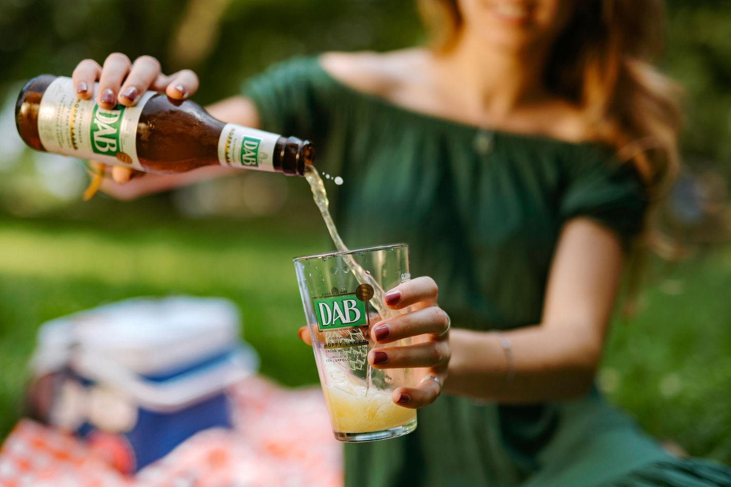 A hand pours a craft beer into a glass at an outdoor gathering with green foliage in the background.
