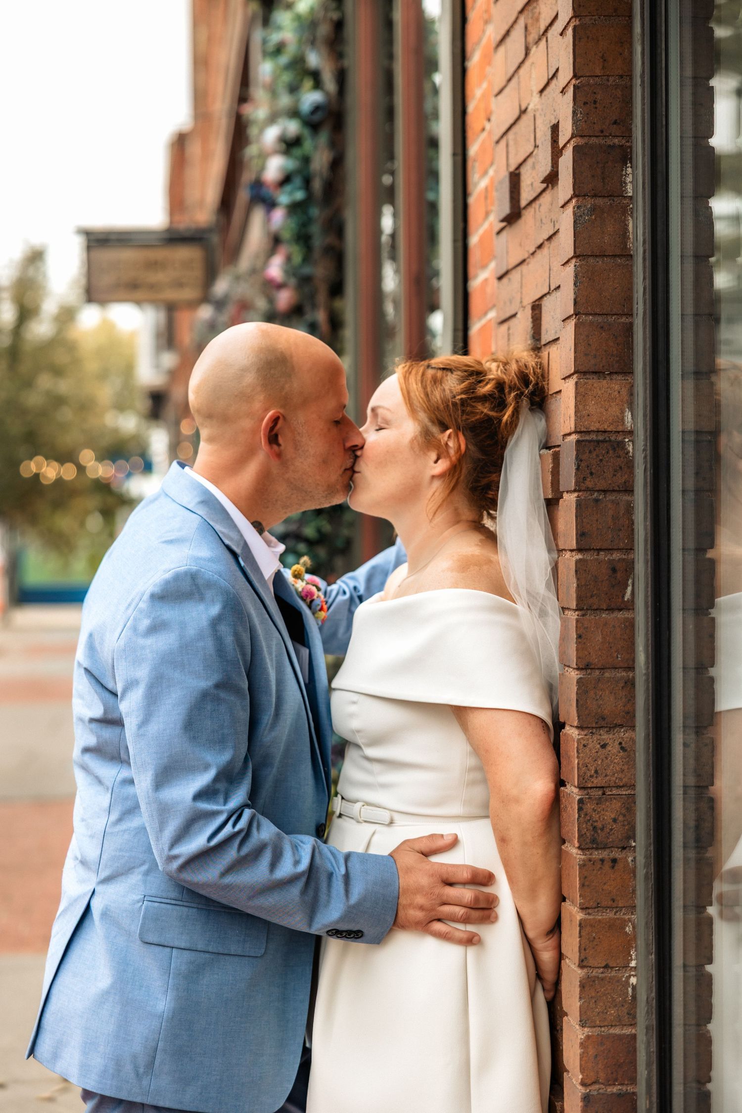 A couple in a blue suit and white dress share a playful moment next to an old brick bldg. Metro Detroit Wedding Content Creator