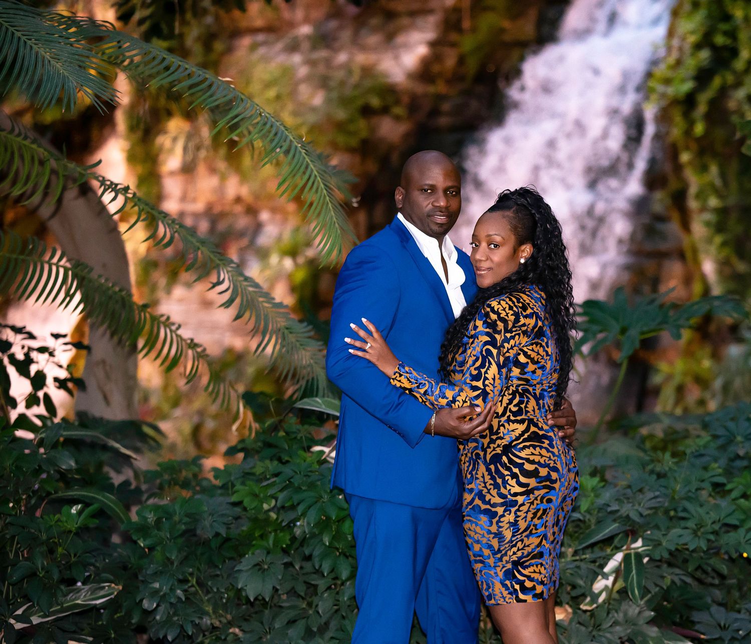 A couple in coordinated blue and leopard print outfits pose together near a scenic waterfall in a tropical setting.
