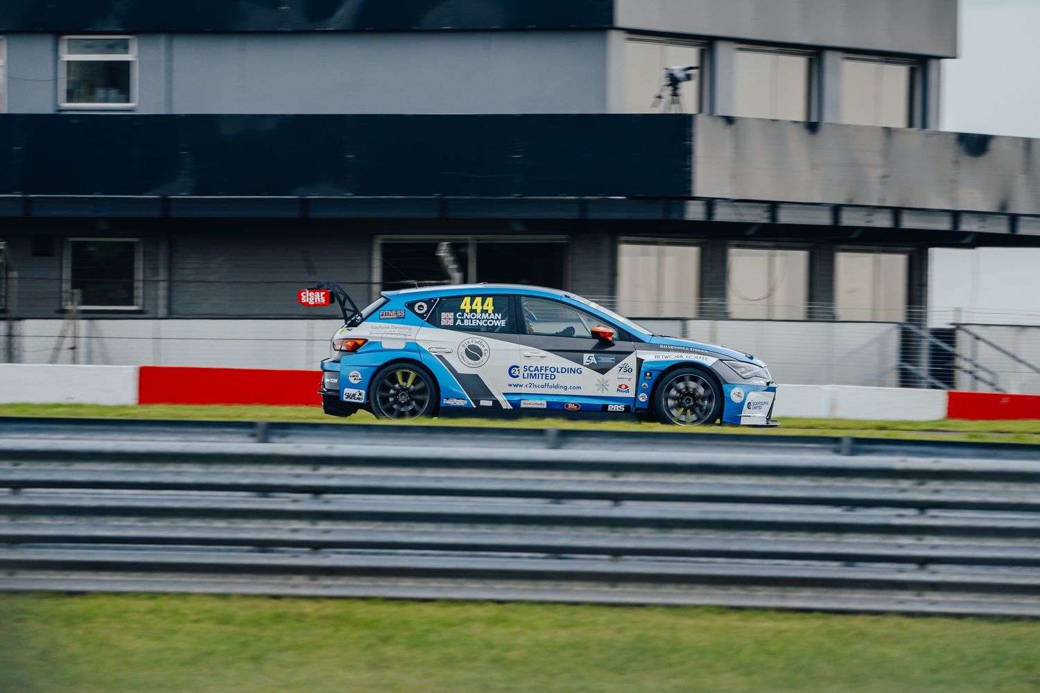 A blue racing car speeds along a racetrack with grandstands and barriers visible in the background.