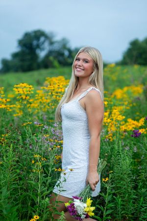 A woman in a white lace dress poses in a vibrant field of yellow and purple wildflowers on a summer day.