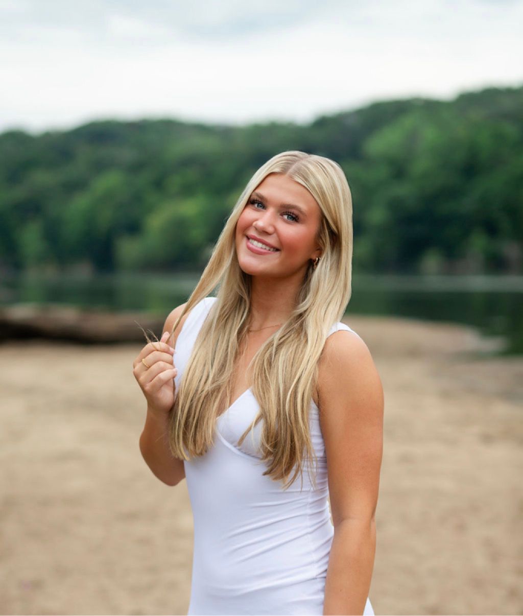A person in a white tank top smiles while standing near a lake with green hills in the background.