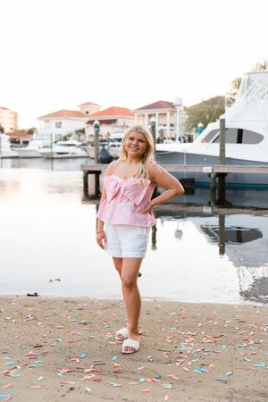 A child plays on a beach with colorful confetti scattered on the sand near a marina with boats in the background.