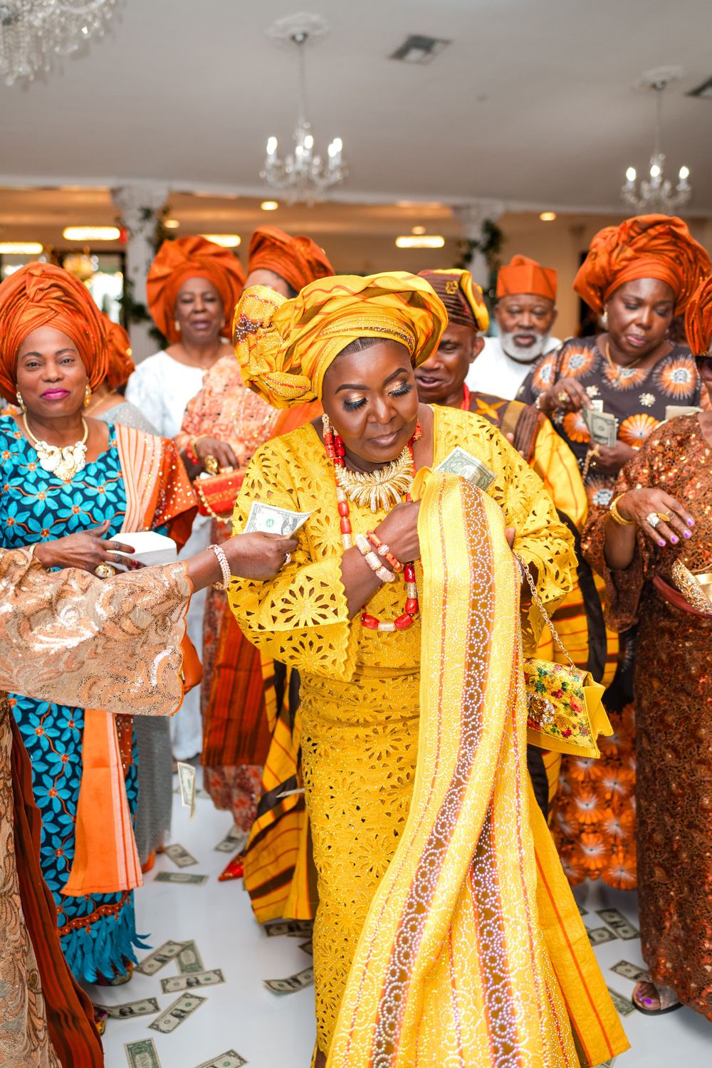 A group celebrates at a Nigerian wedding ceremony wearing vibrant yellow and orange traditional attire and gele headwraps.