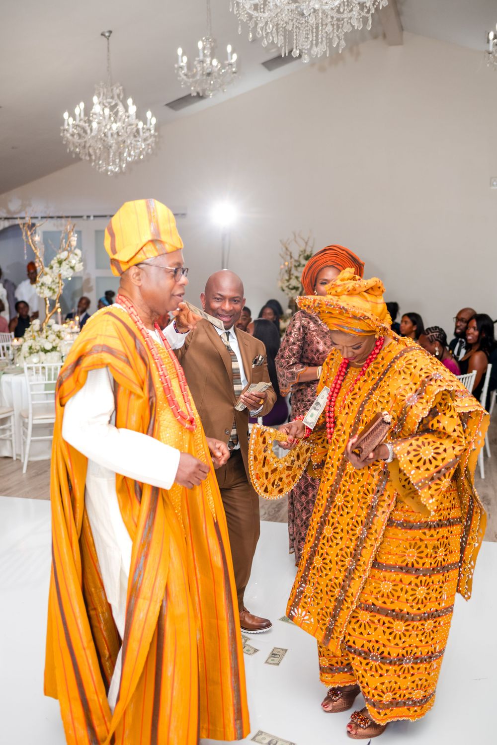 Traditional African wedding celebration with guests wearing vibrant orange and leopard print attire dancing in an elegant venue.