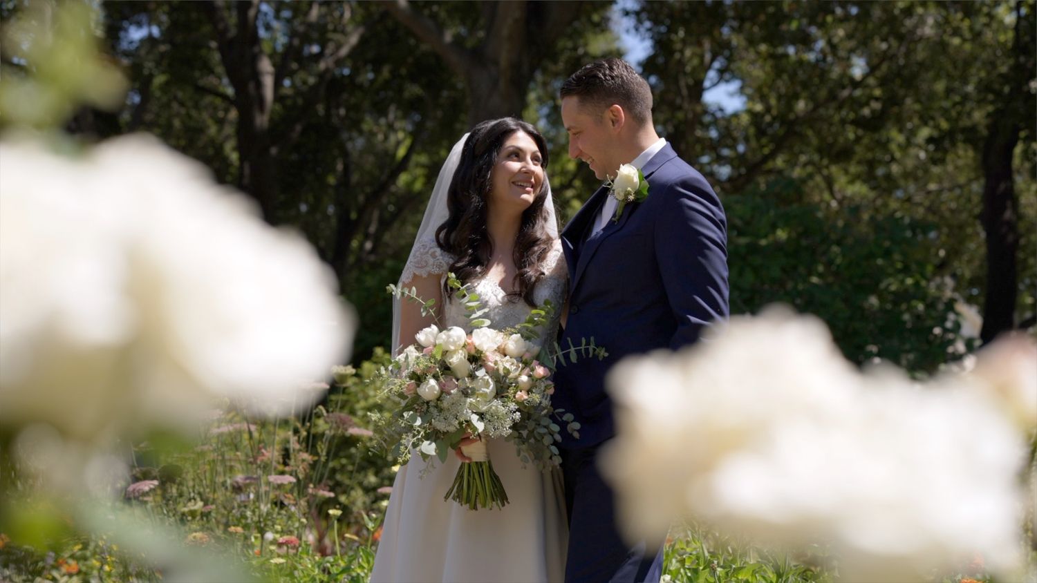 A couple embraces in a romantic garden setting surrounded by blooming white flowers and lush greenery.