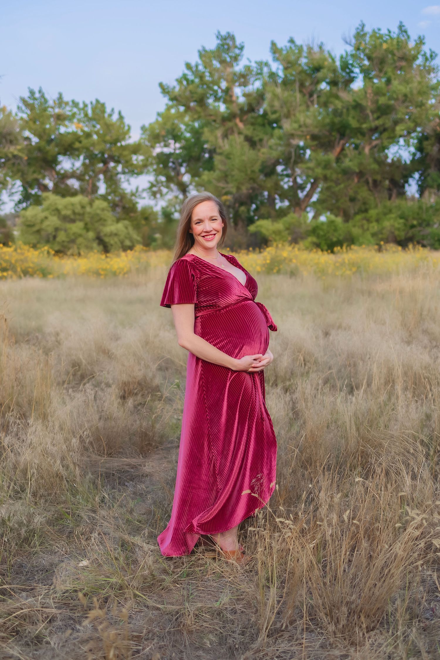 Side side of a pregnant woman in a long burgundy dress.  She is outside and cradling her bump