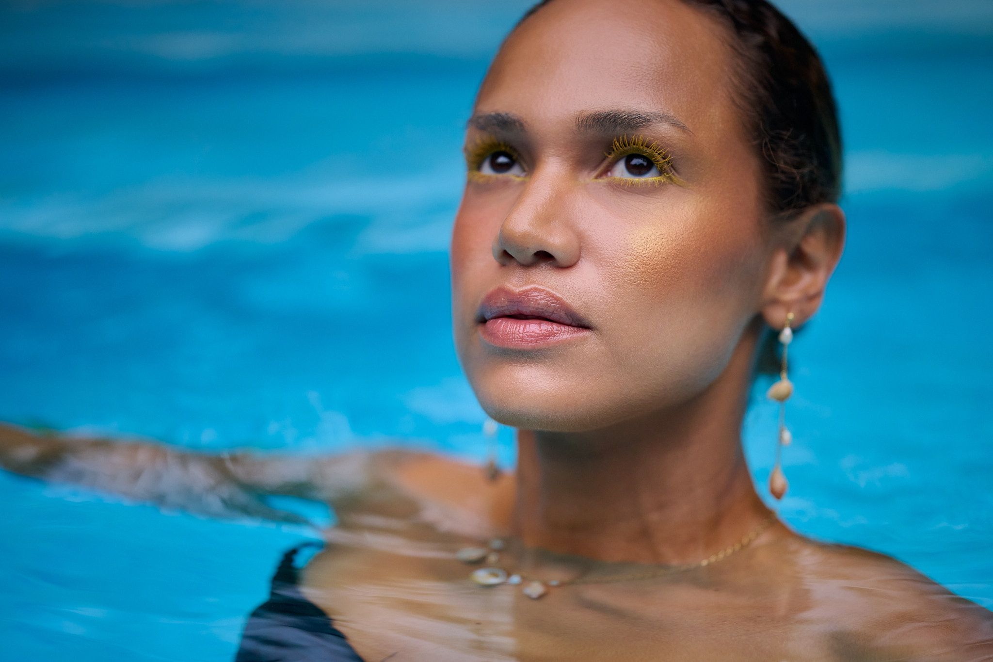 A woman emerges from bright blue pool water with a serene expression and glistening skin.