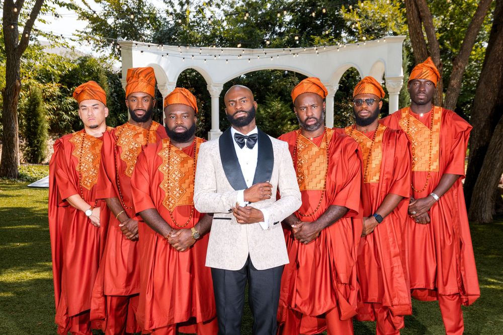 A wedding party poses outdoors with the groom in a white suit jacket and groomsmen wearing bright orange robes.