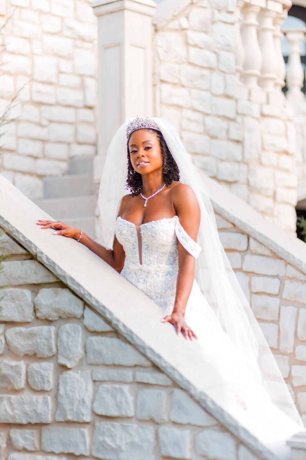 A bride in a stunning white gown and tiara poses gracefully on stone stairs.