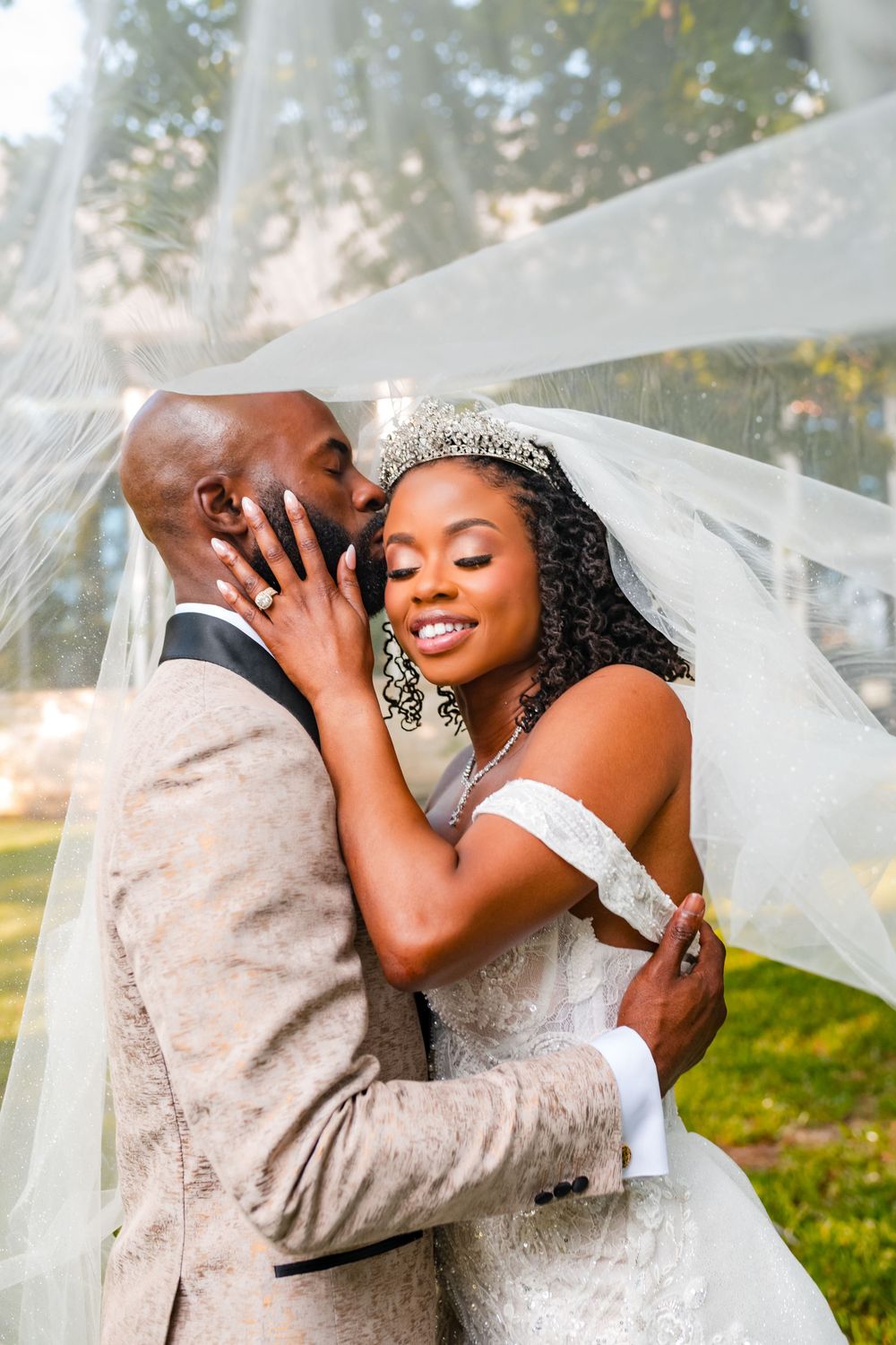 A joyful wedding couple embraces under a flowing veil in an outdoor garden setting.