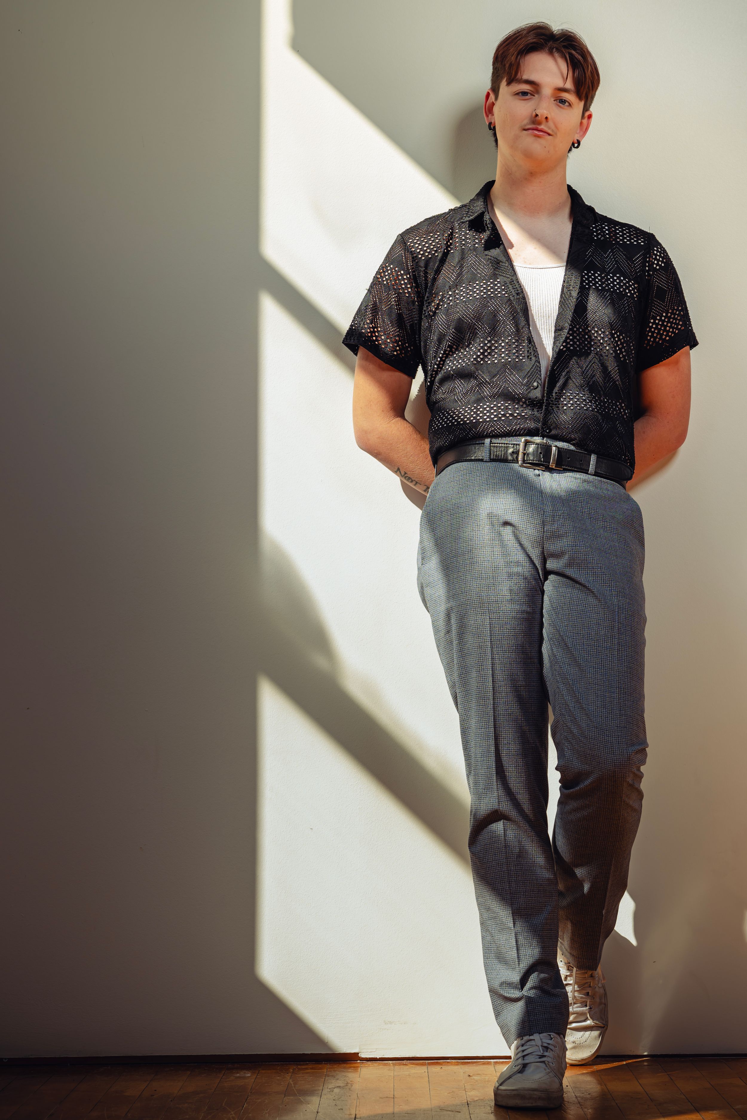 A person in a black patterned short-sleeve shirt and gray pants poses against a white wall with dramatic shadows.