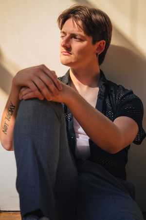 A person in a dark sweater sits against a beige wall while showing their forearm tattoo in dramatic lighting.
