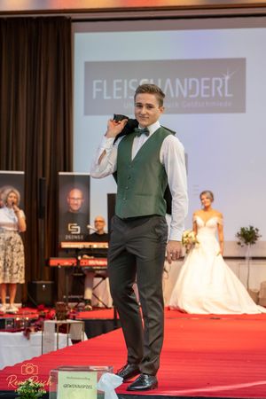 Male model presents green formal wear on red carpet runway with bridal show backdrop.