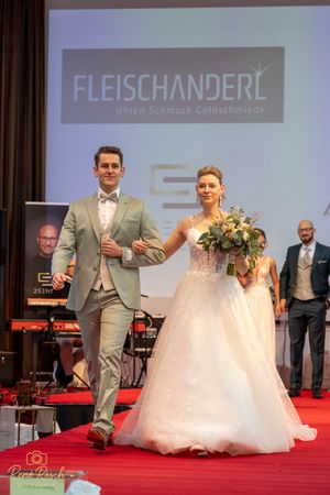 Models showcase elegant wedding attire on a red carpet runway with Fleischanderl branding displayed in the background.